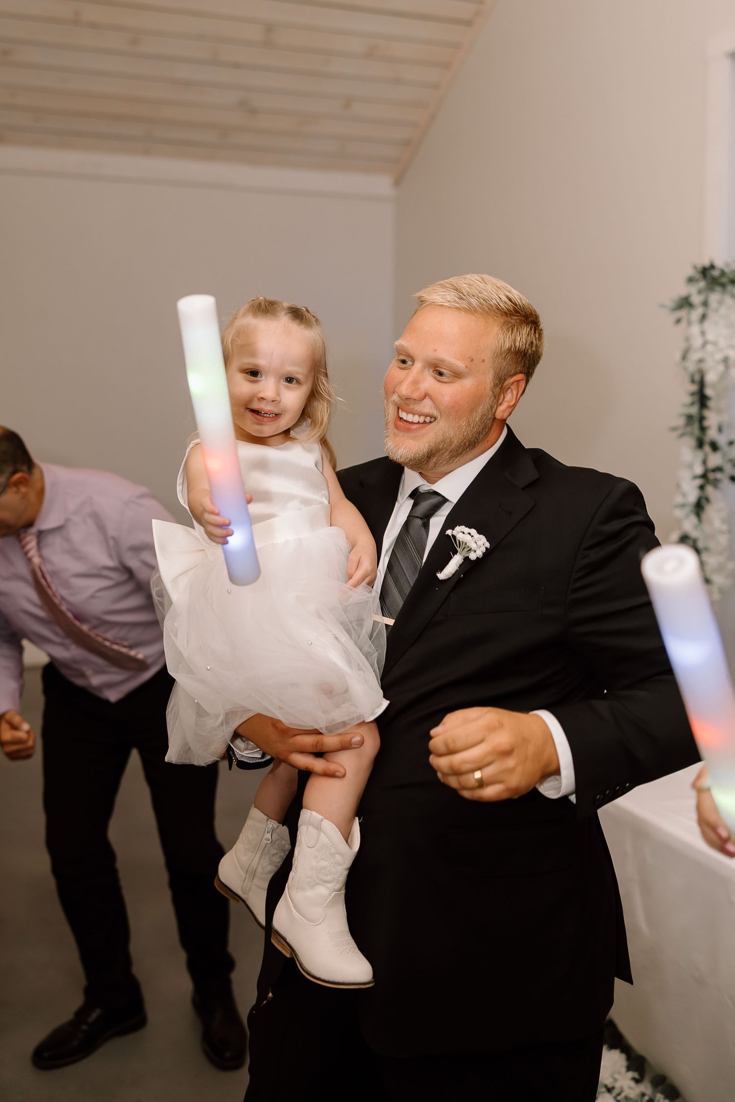 A person in a black suit holds a small child in white during what appears to be a wedding celebration.