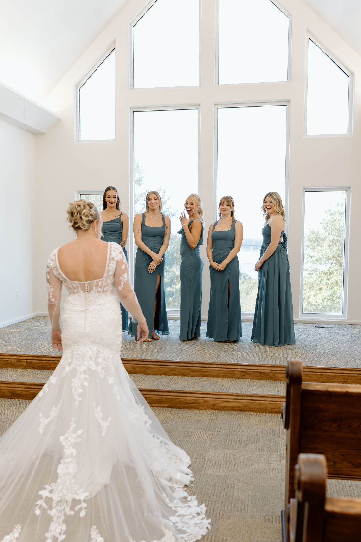 Bridesmaids in matching teal dresses stand in a row inside a bright church venue with white walls and wooden pews.