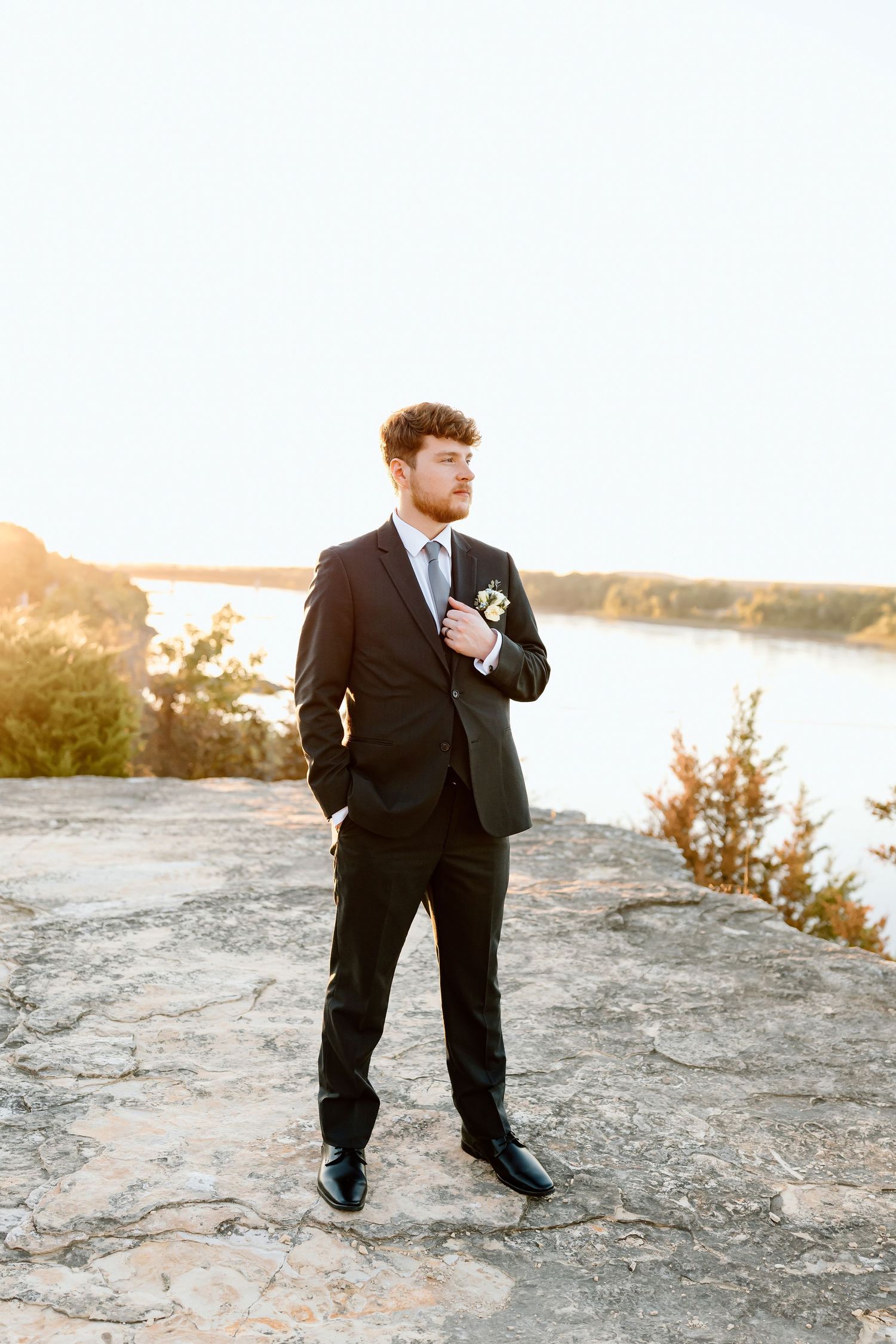 A person in a black suit stands on rocky terrain at sunset, creating a silhouetted figure against the golden sky.