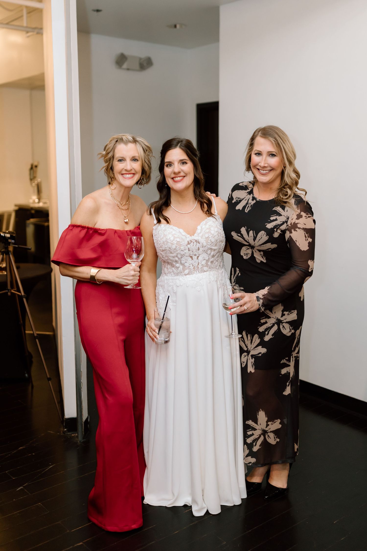 Three women pose together at a formal event wearing elegant dresses in red, white and black floral patterns.