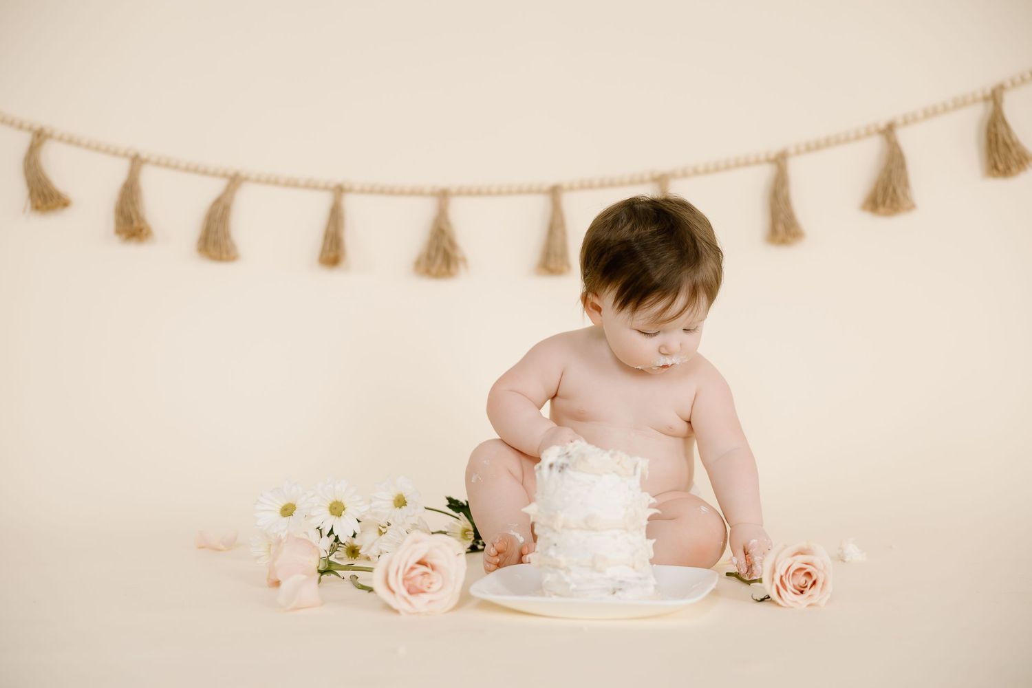 Baby in diaper examining white cake with pink roses against tassel-decorated backdrop.