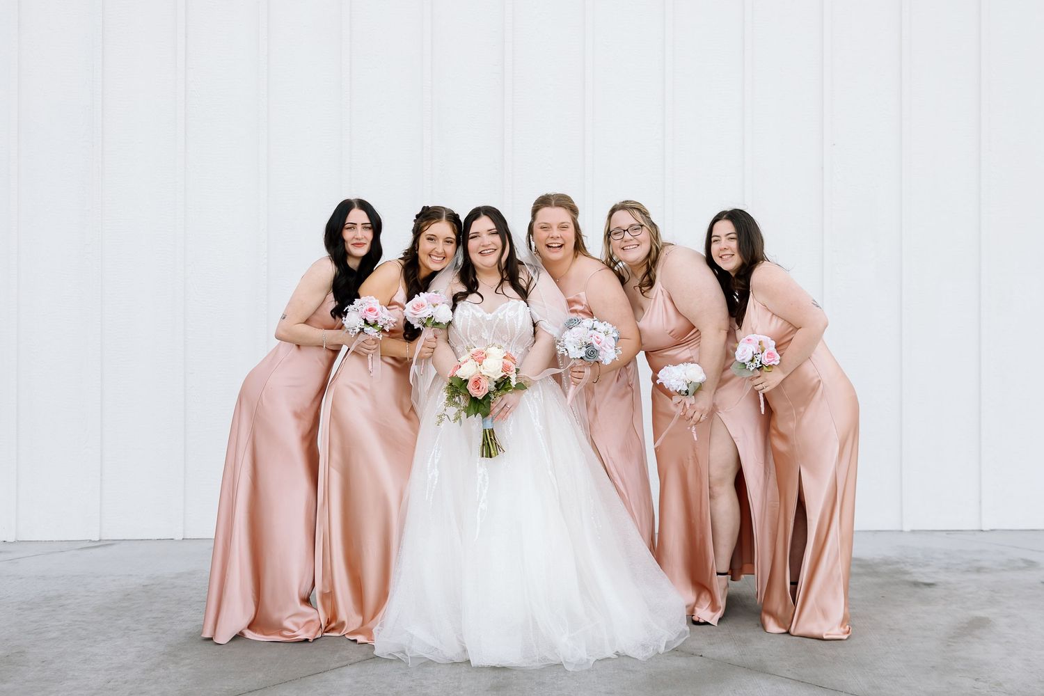 Wedding party with bridesmaids in blush pink satin gowns posing against white wall.