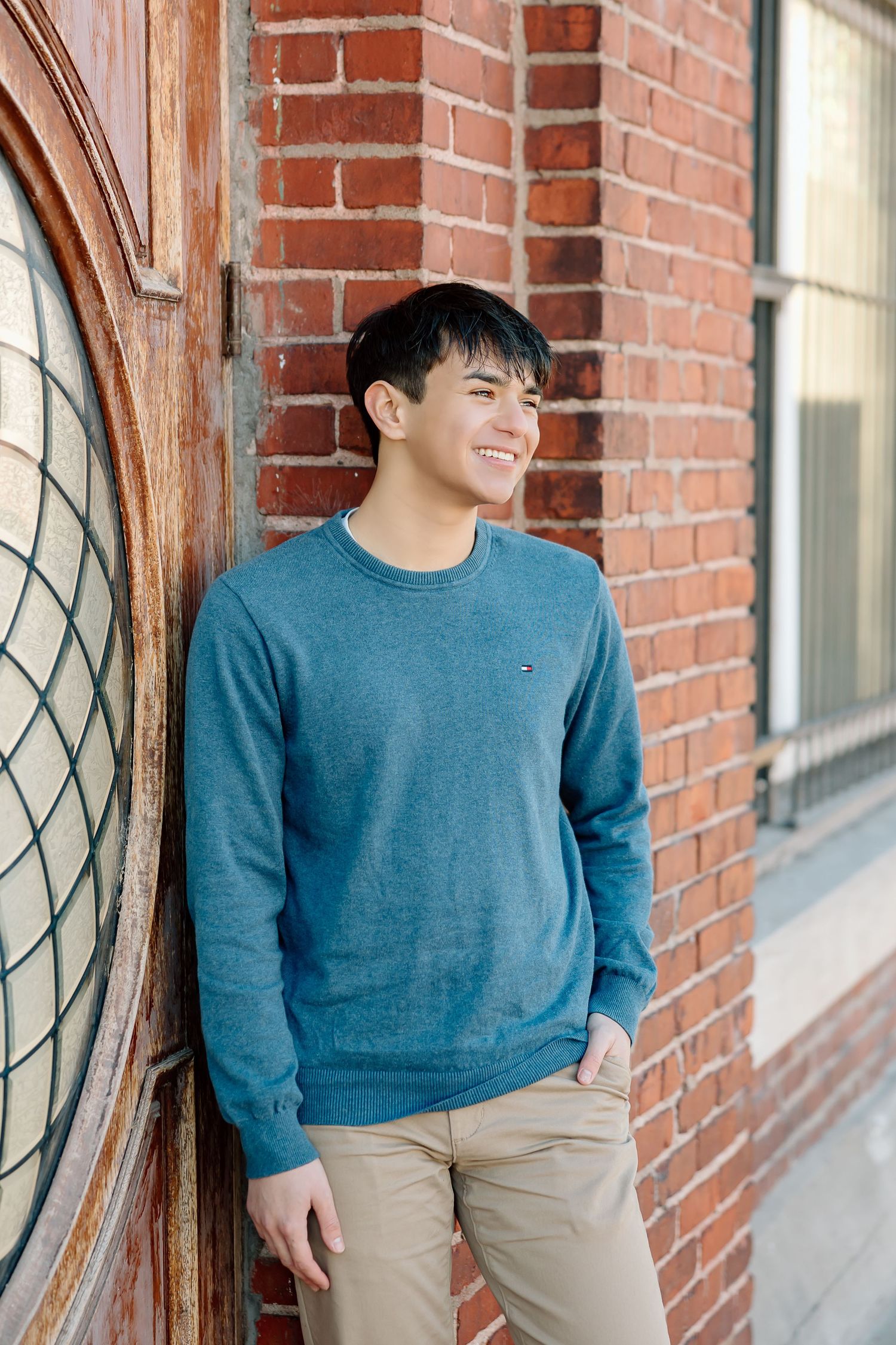 Subject in blue sweater smiling against red brick wall with window.