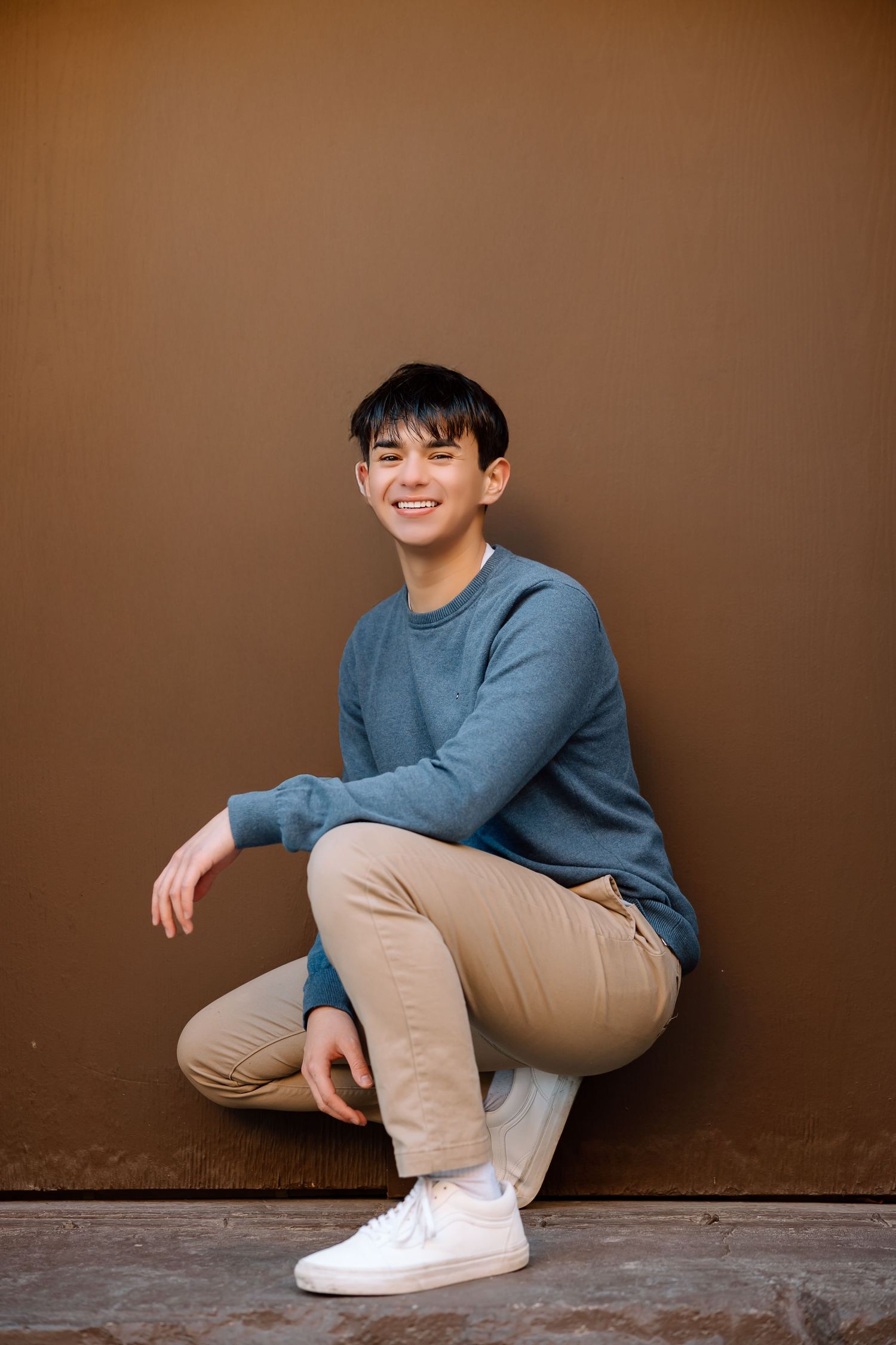 Portrait against brown background wearing blue sweater and khaki pants in crouching pose.