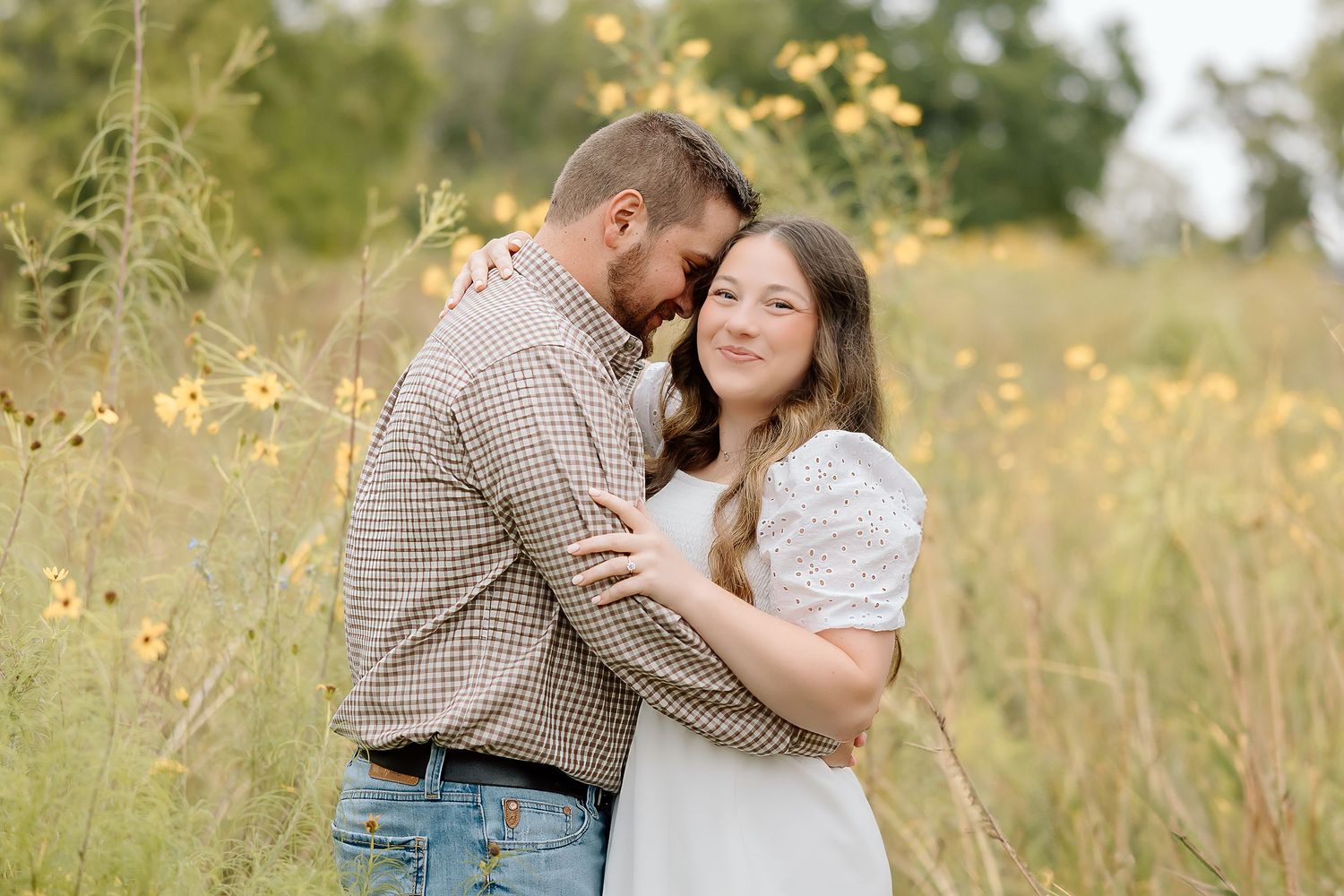 A couple in casual attire shares a loving moment while standing in a sun-drenched meadow with yellow flowers.