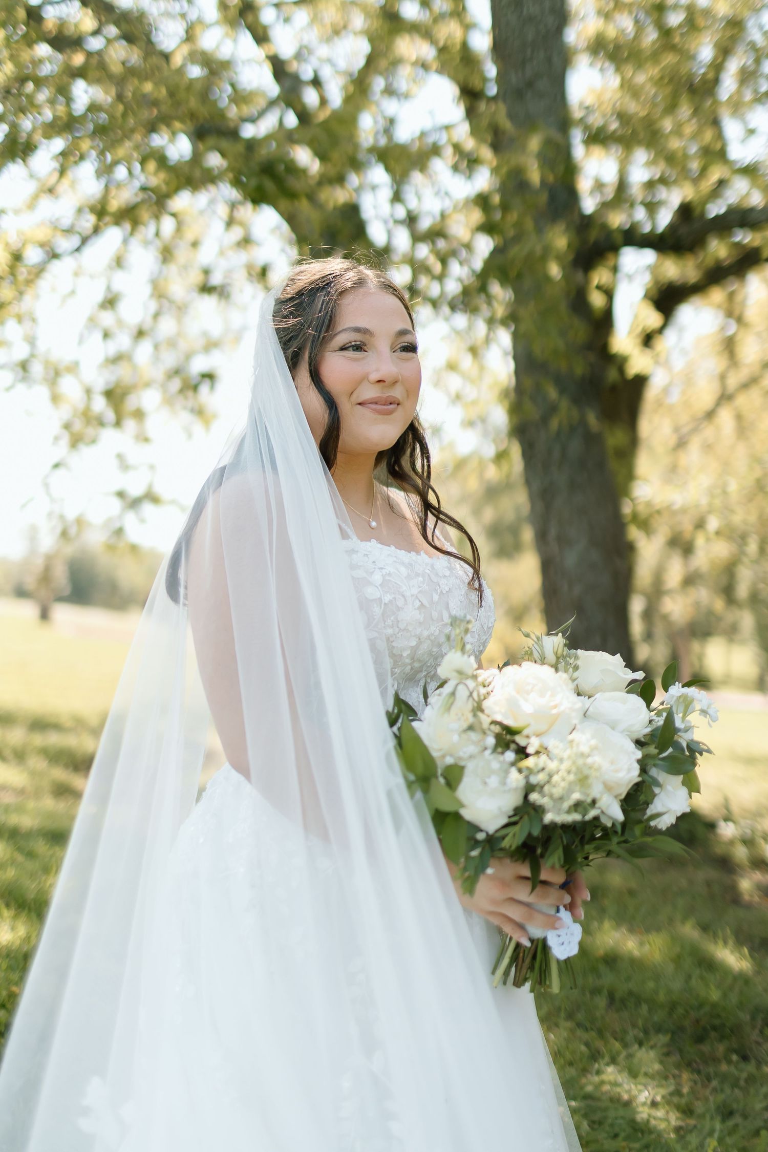 A bride in a white wedding gown and veil poses outdoors among trees holding a white floral bouquet.