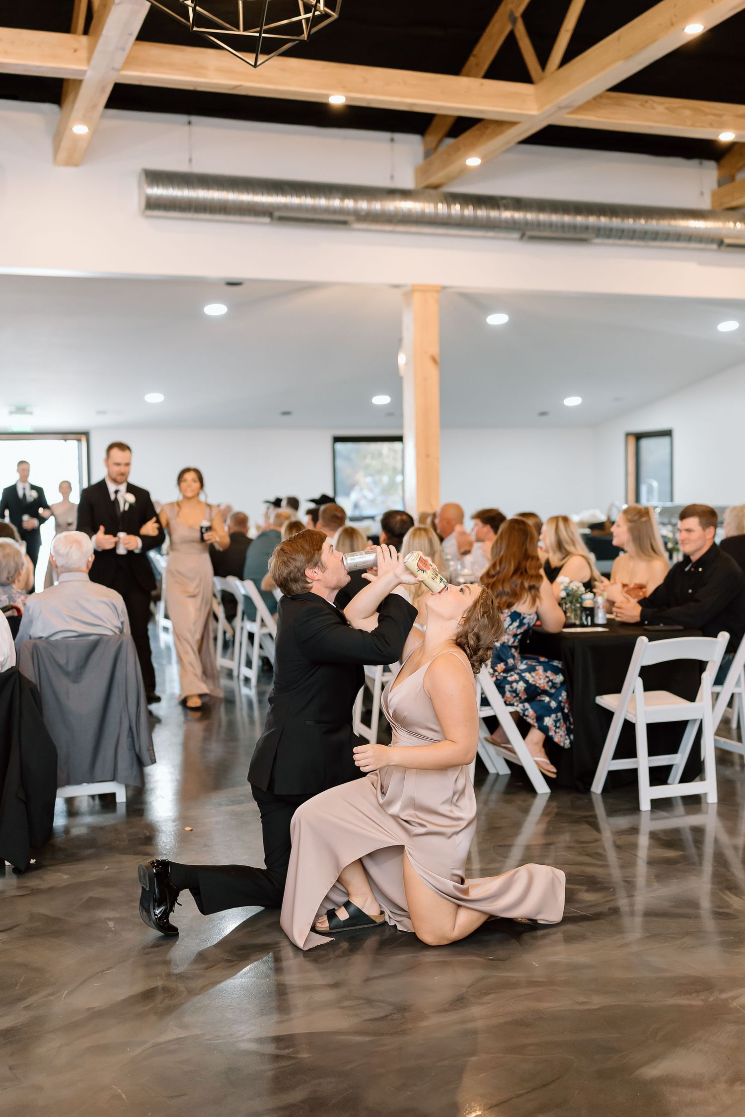 Wedding guests enjoying a lively dance party in an industrial-style reception venue with exposed beams and white chairs.