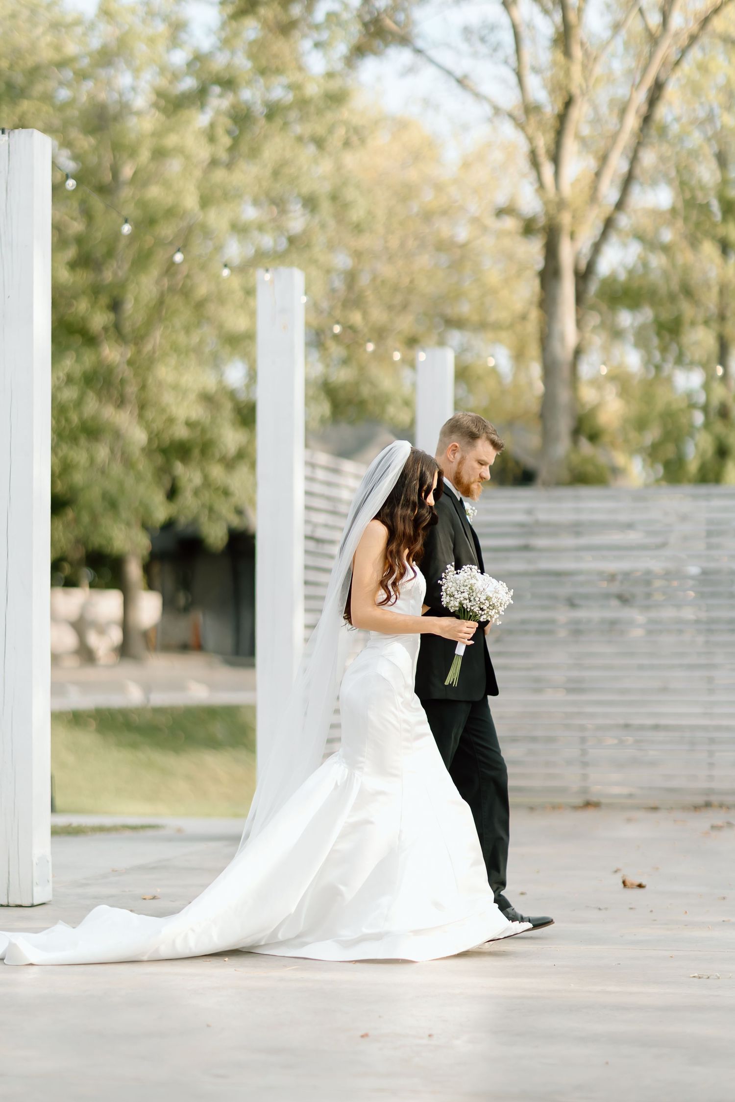 A couple shares an intimate moment in their wedding attire against white architectural columns and autumn trees.