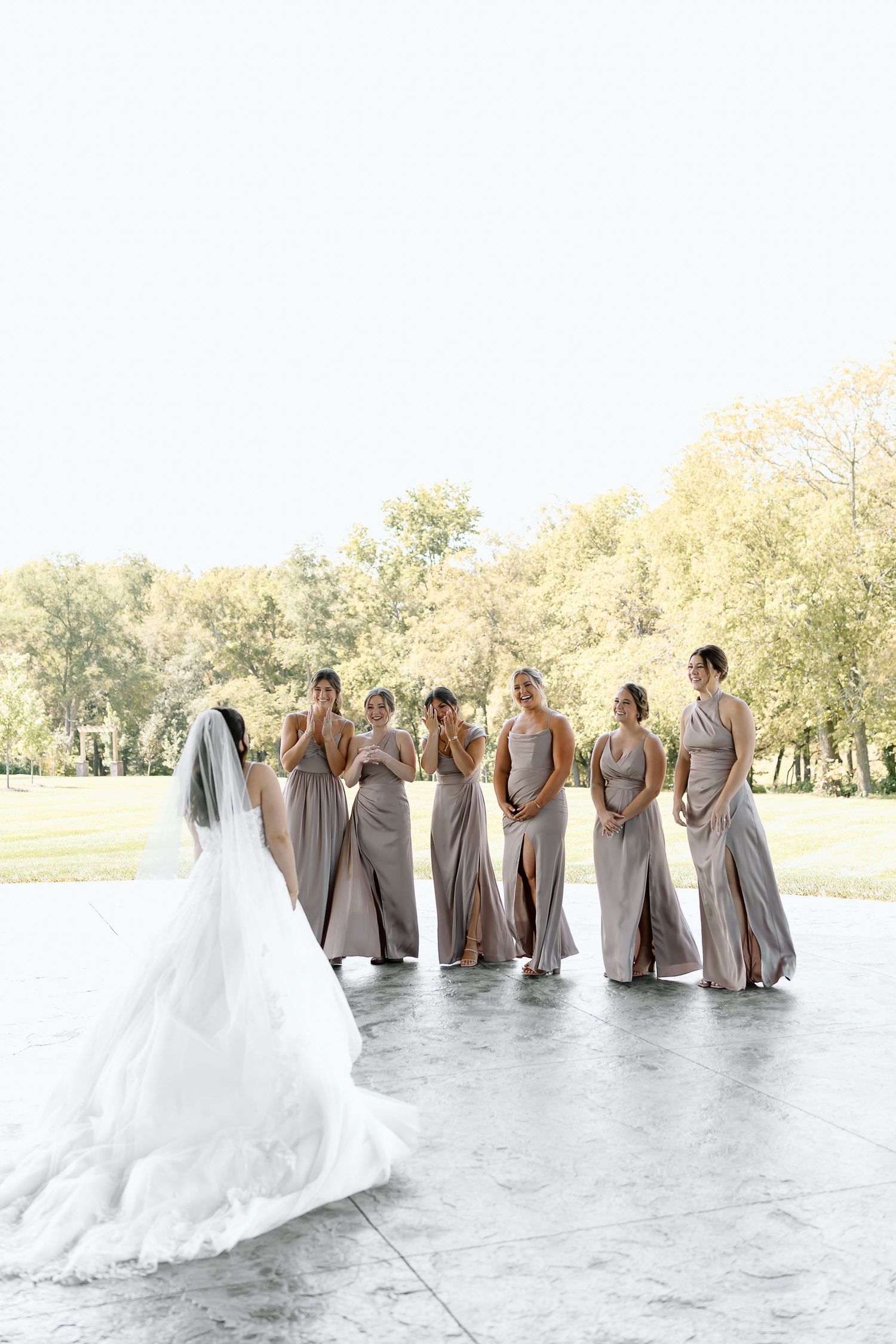Bridesmaids in flowing gray dresses react with joy to bride's stunning white wedding gown on outdoor patio.