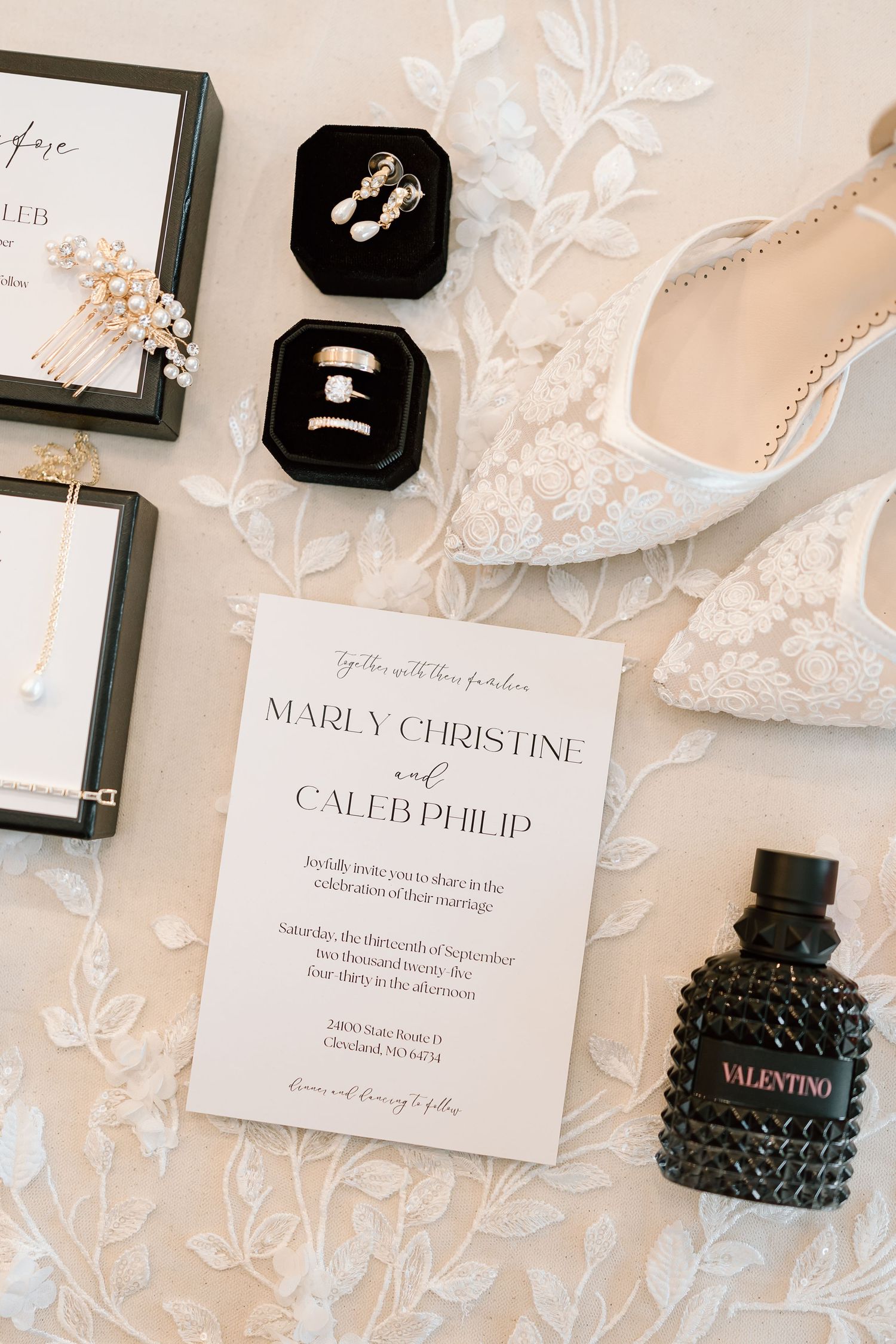 Elegant wedding invitations and rings displayed on white lace with delicate floral details and black velvet ring boxes.
