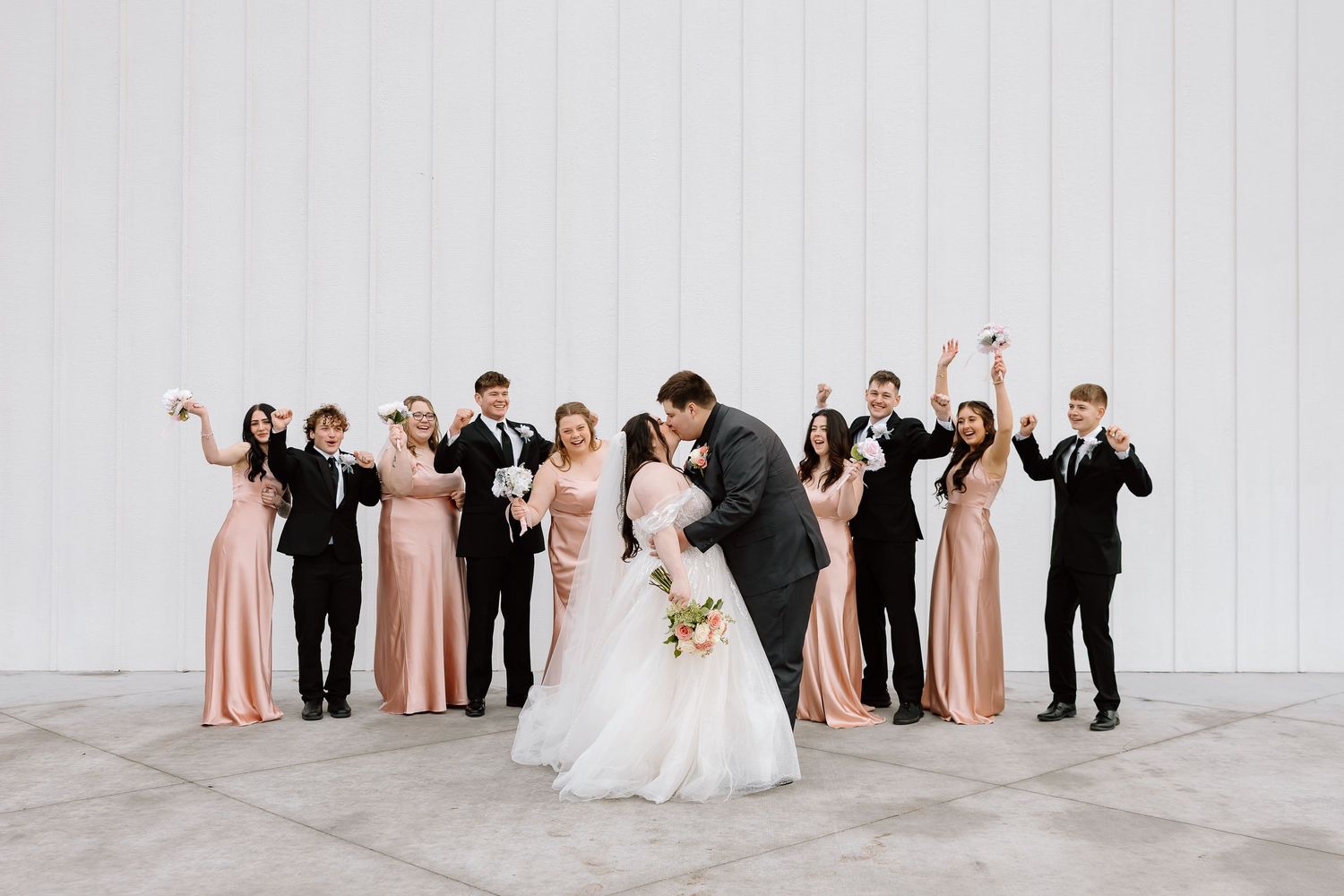 Wedding party in formal attire poses together in line against white wall wearing black and blush pink colors.