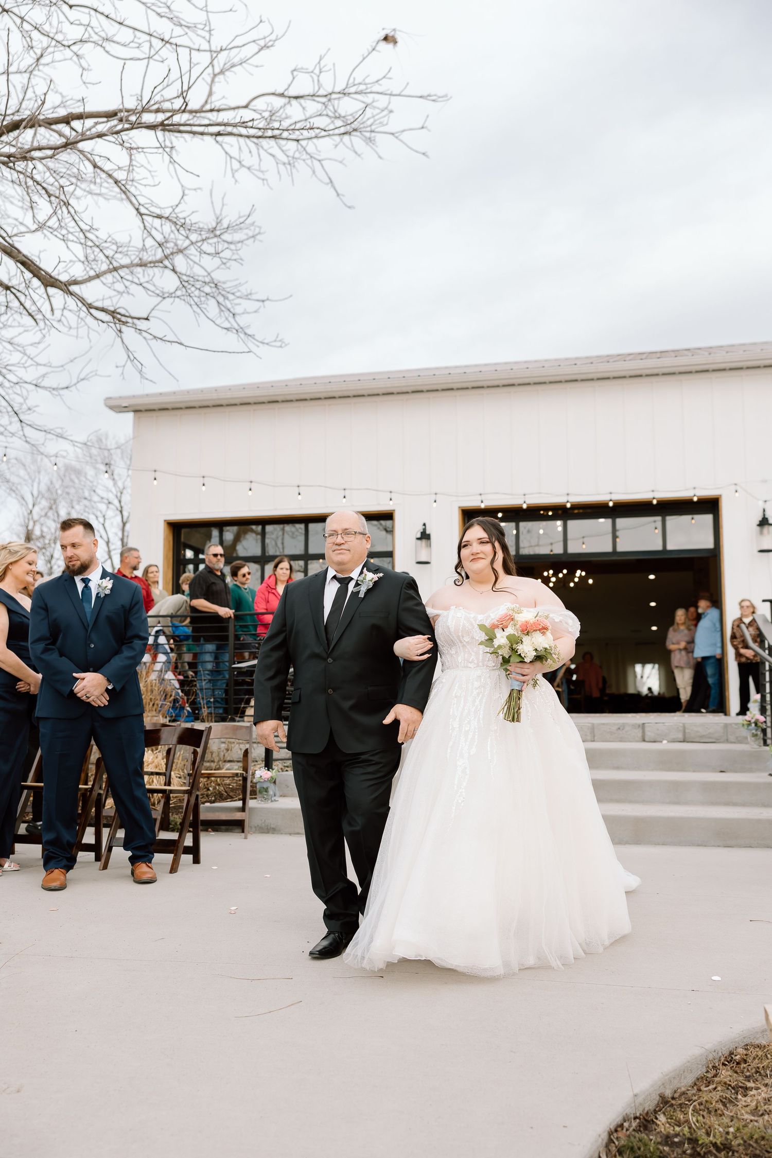 A wedding ceremony takes place outside a modern white venue with guests gathered on a cloudy winter day.