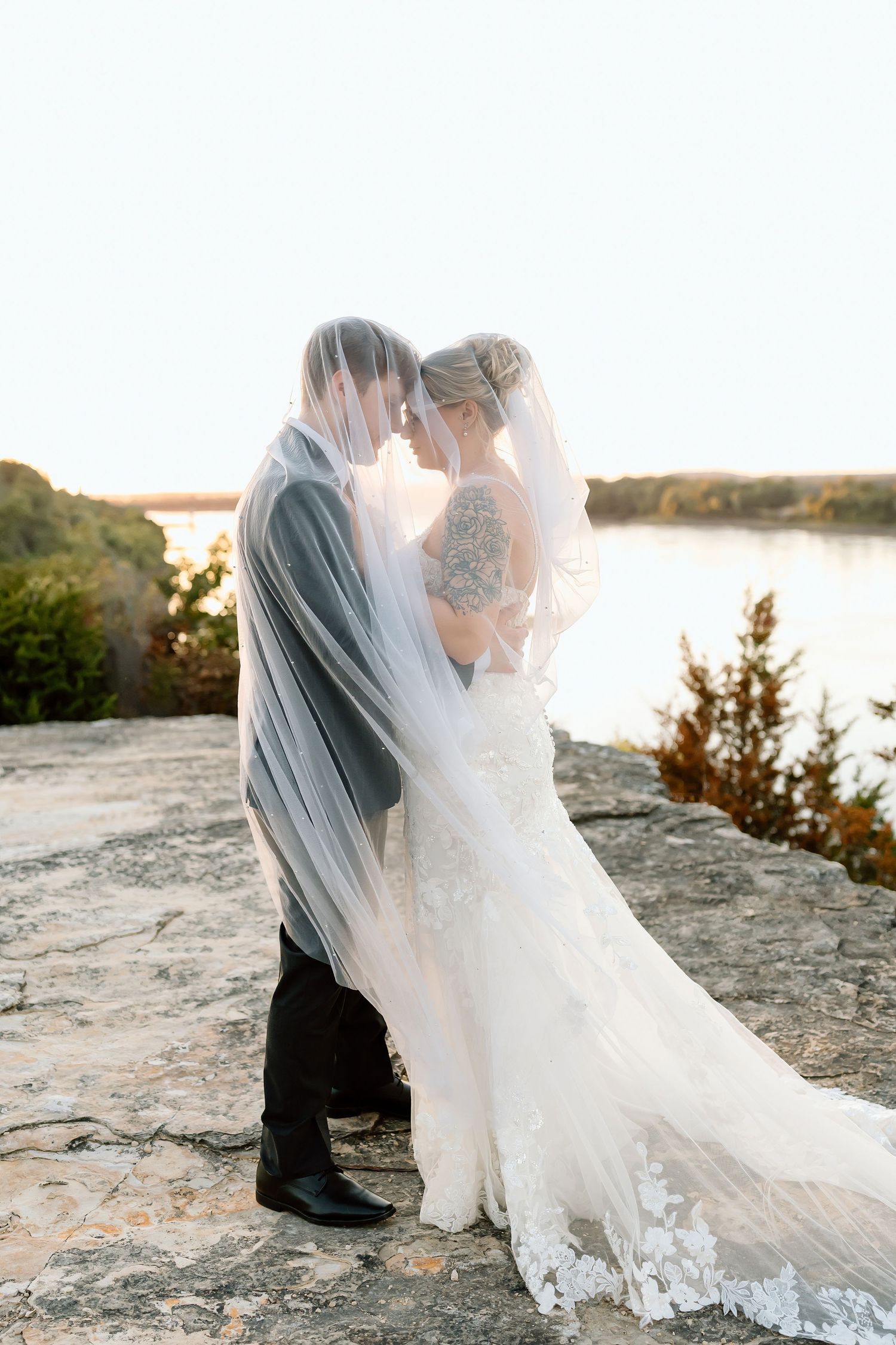 A romantic wedding couple embraces on a cliff overlooking a serene lake at sunset, with flowing veils and lace details.