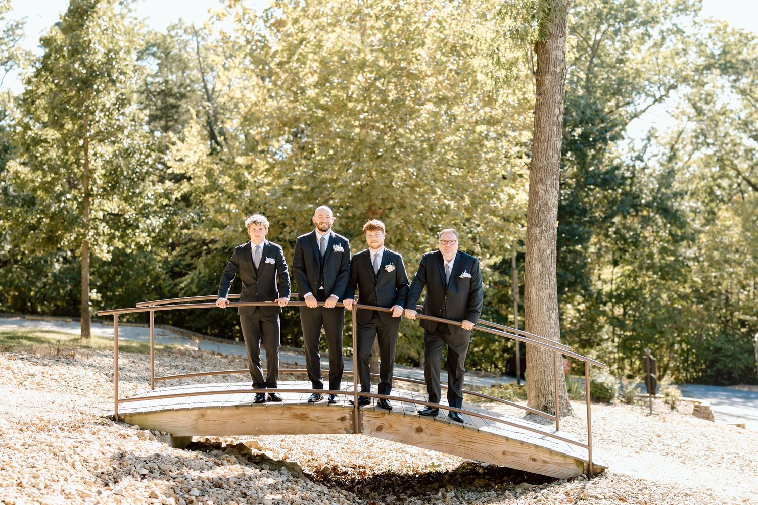 Wedding party members gather on a rustic wooden bridge surrounded by autumn trees in an outdoor setting.