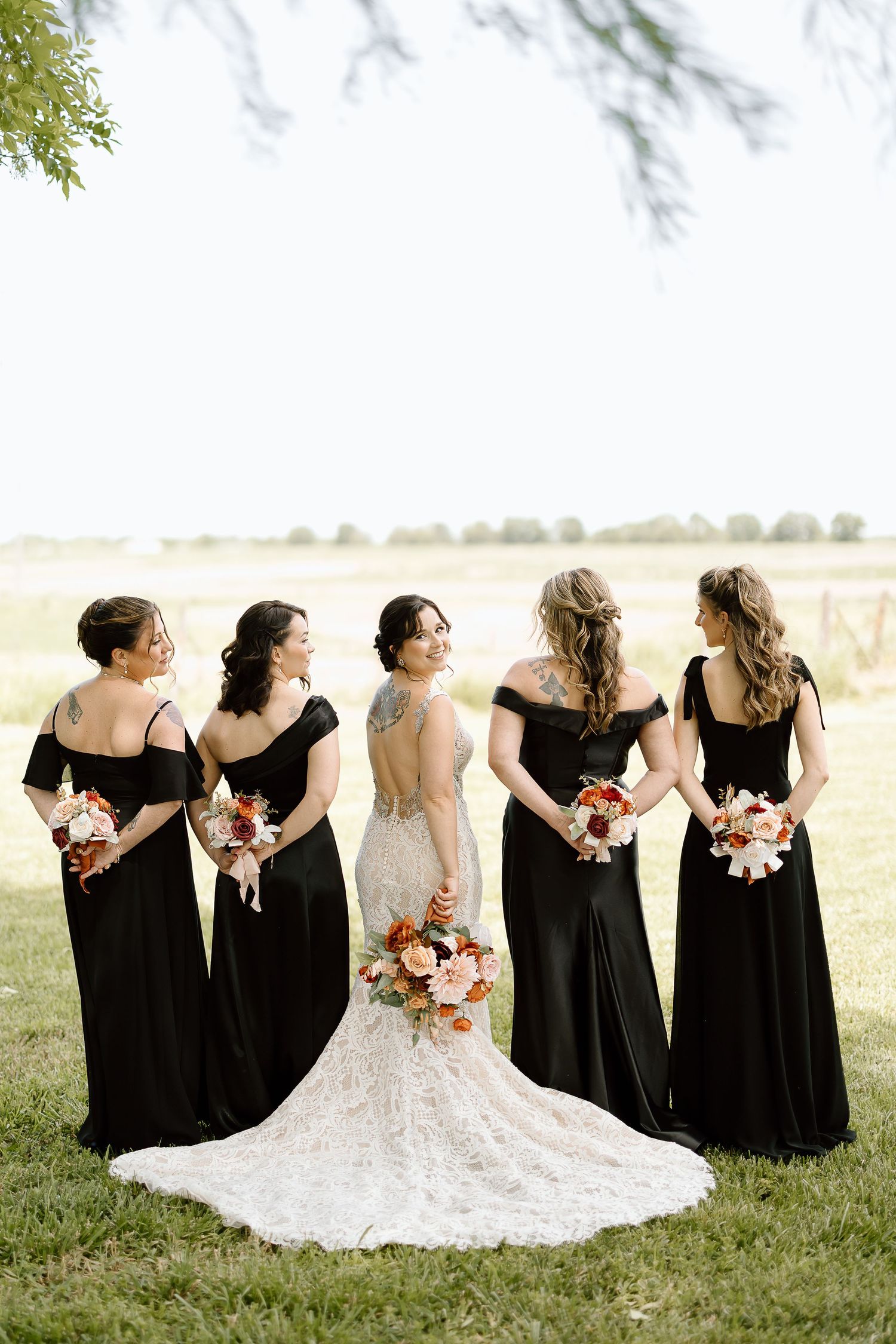 A bride in white lace gown stands with bridesmaids in black dresses holding bouquets in a sunny outdoor field.