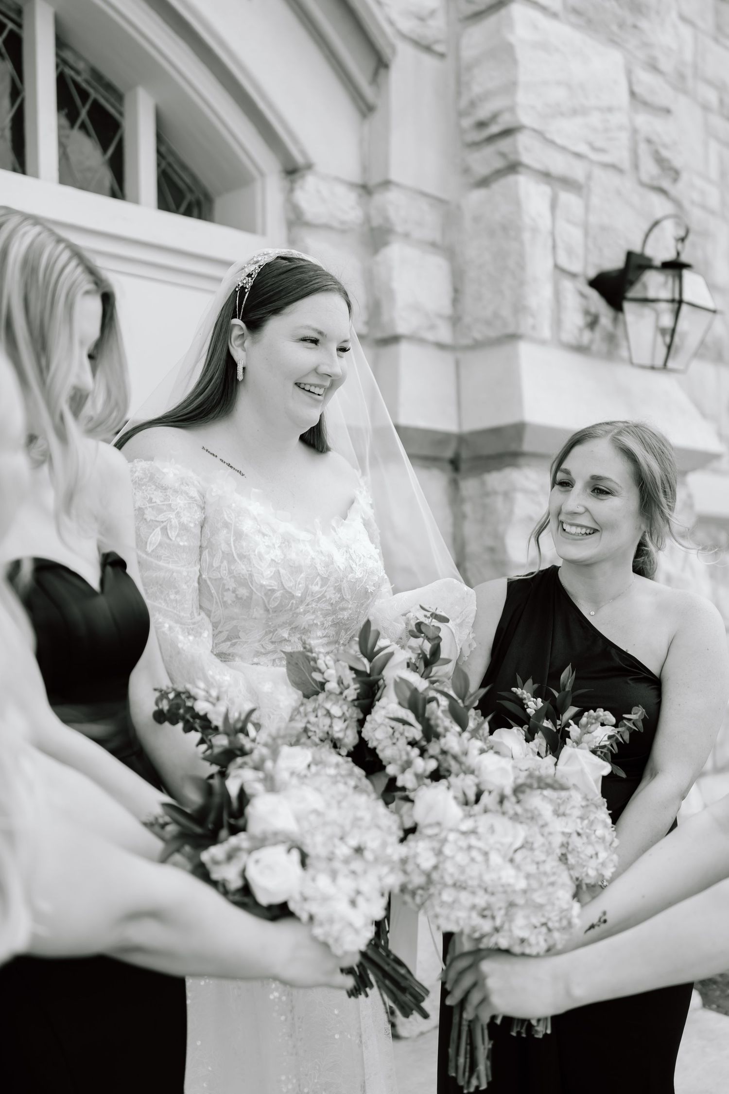 Black and white photo of a smiling bride in lace gown and veil with bridesmaids in black dresses holding floral bouquets.