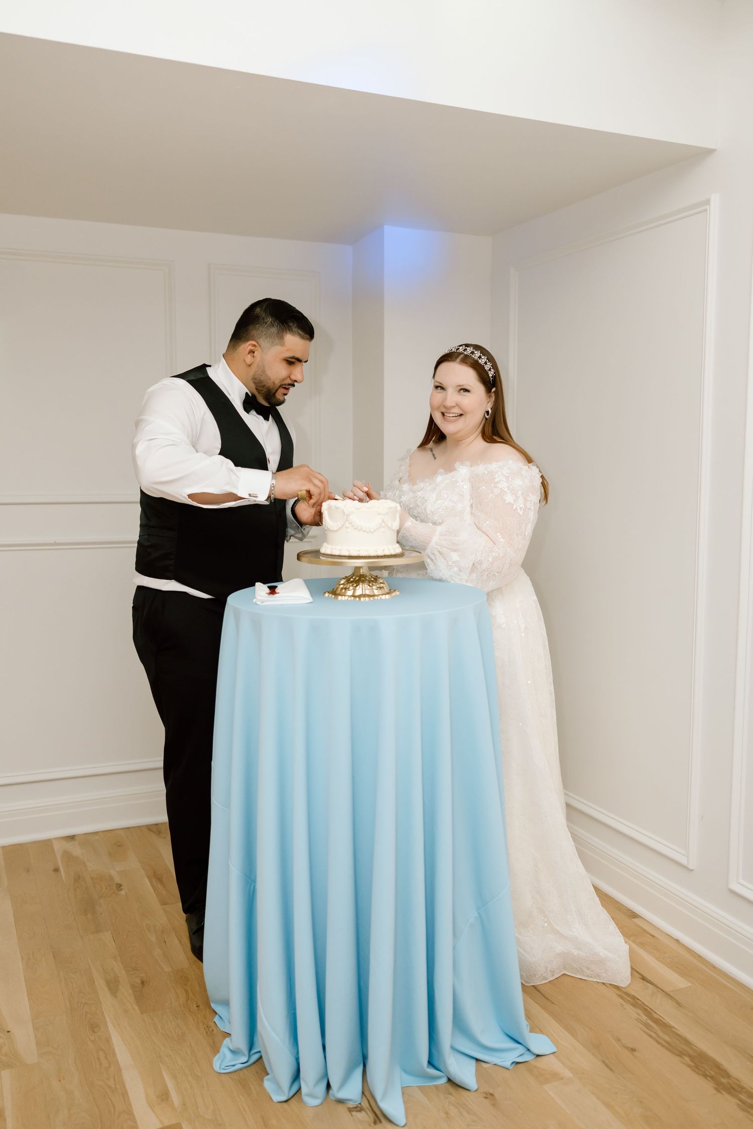 A bride in a white lace gown and groom cut a white wedding cake on a light blue draped table.