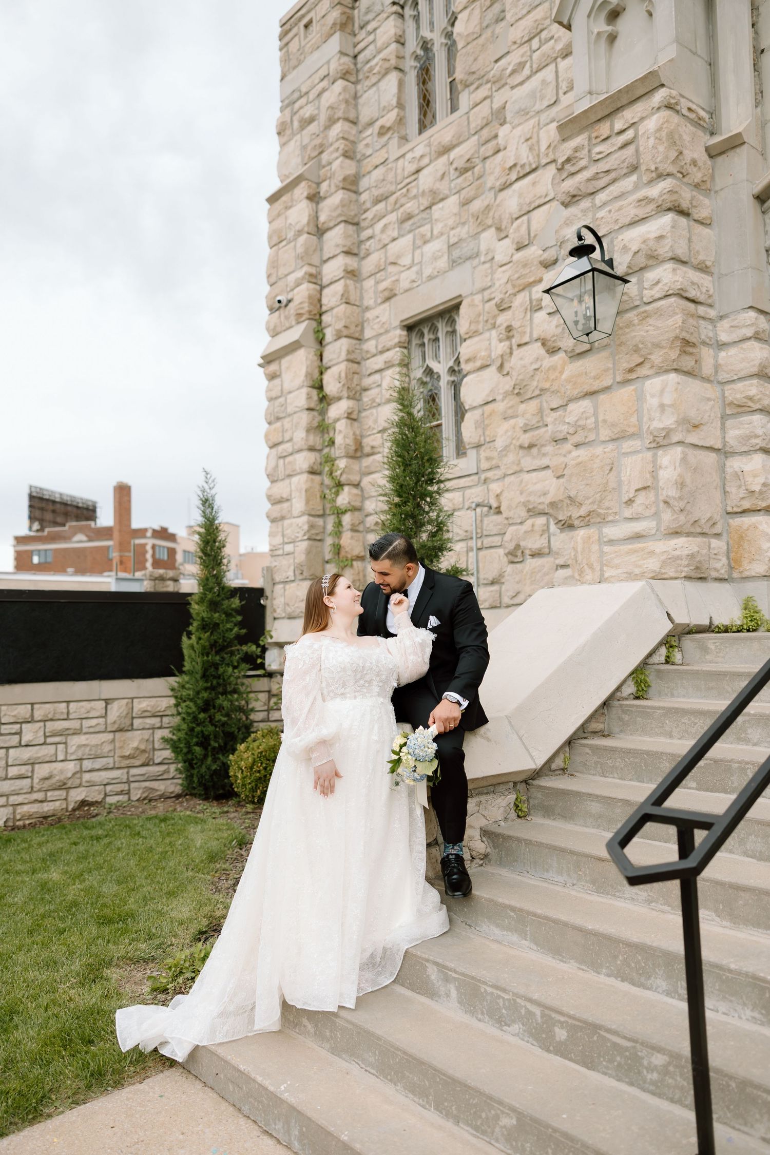 Bride and groom share a romantic moment on stone church steps, bride in white gown holding bouquet.