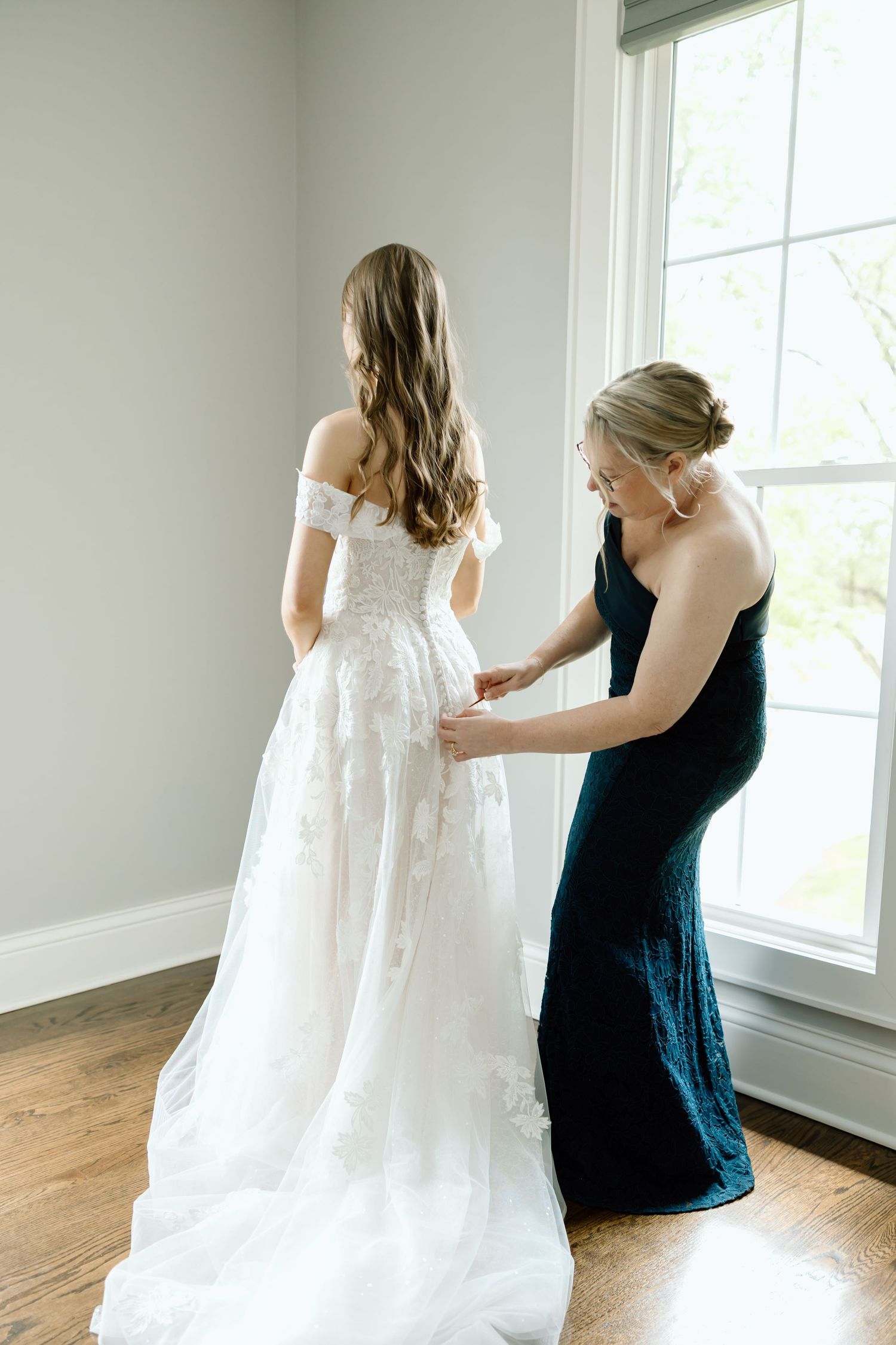 Bride in white lace off-shoulder gown being helped by bridesmaid in navy dress near bright window.