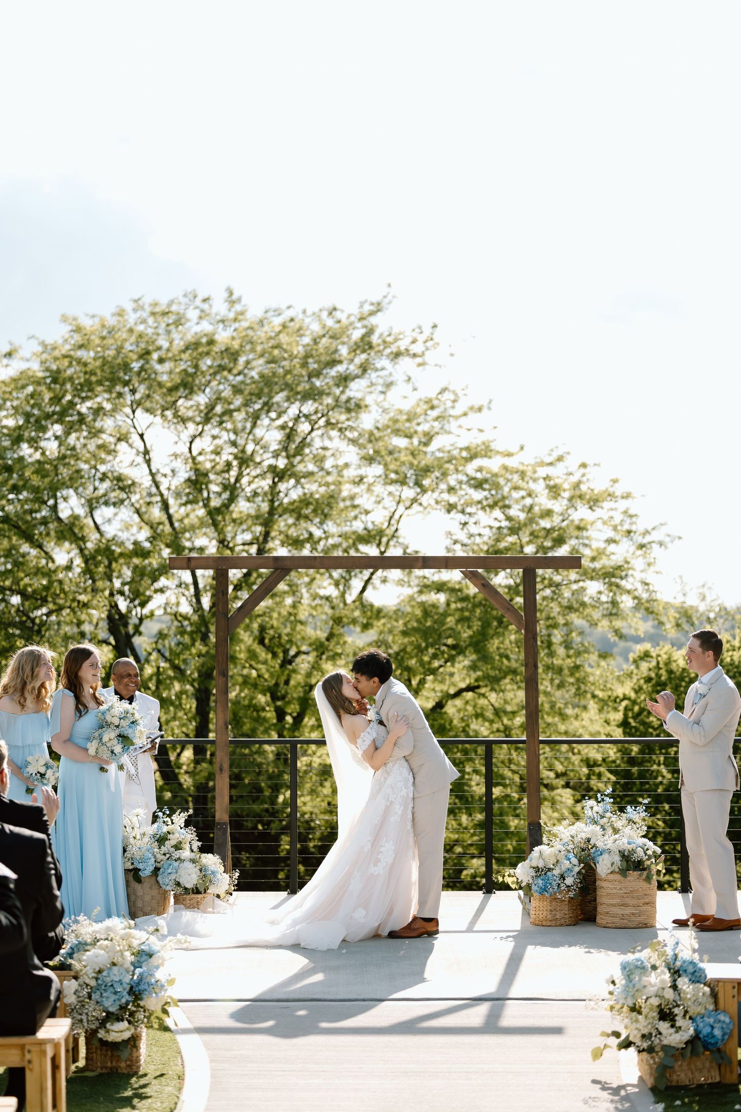 Couple shares first kiss at outdoor wedding ceremony with wooden arch, blue floral arrangements, and bridesmaids in light blue dresses.