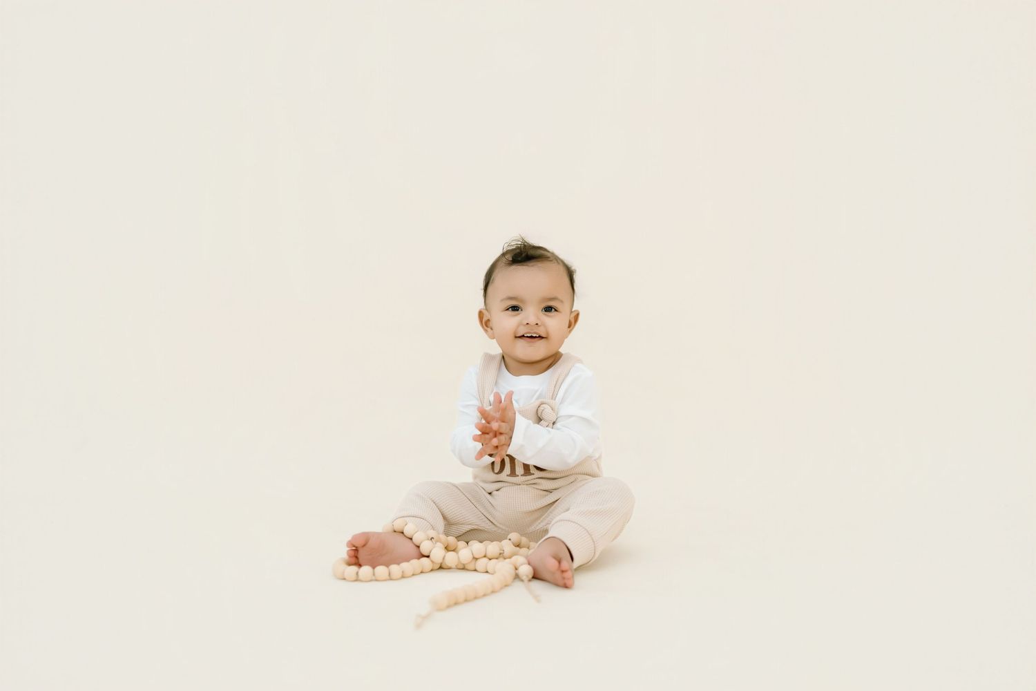 A baby in white onesie sits on a light beige backdrop smiling and looking upward during a professional photo session.