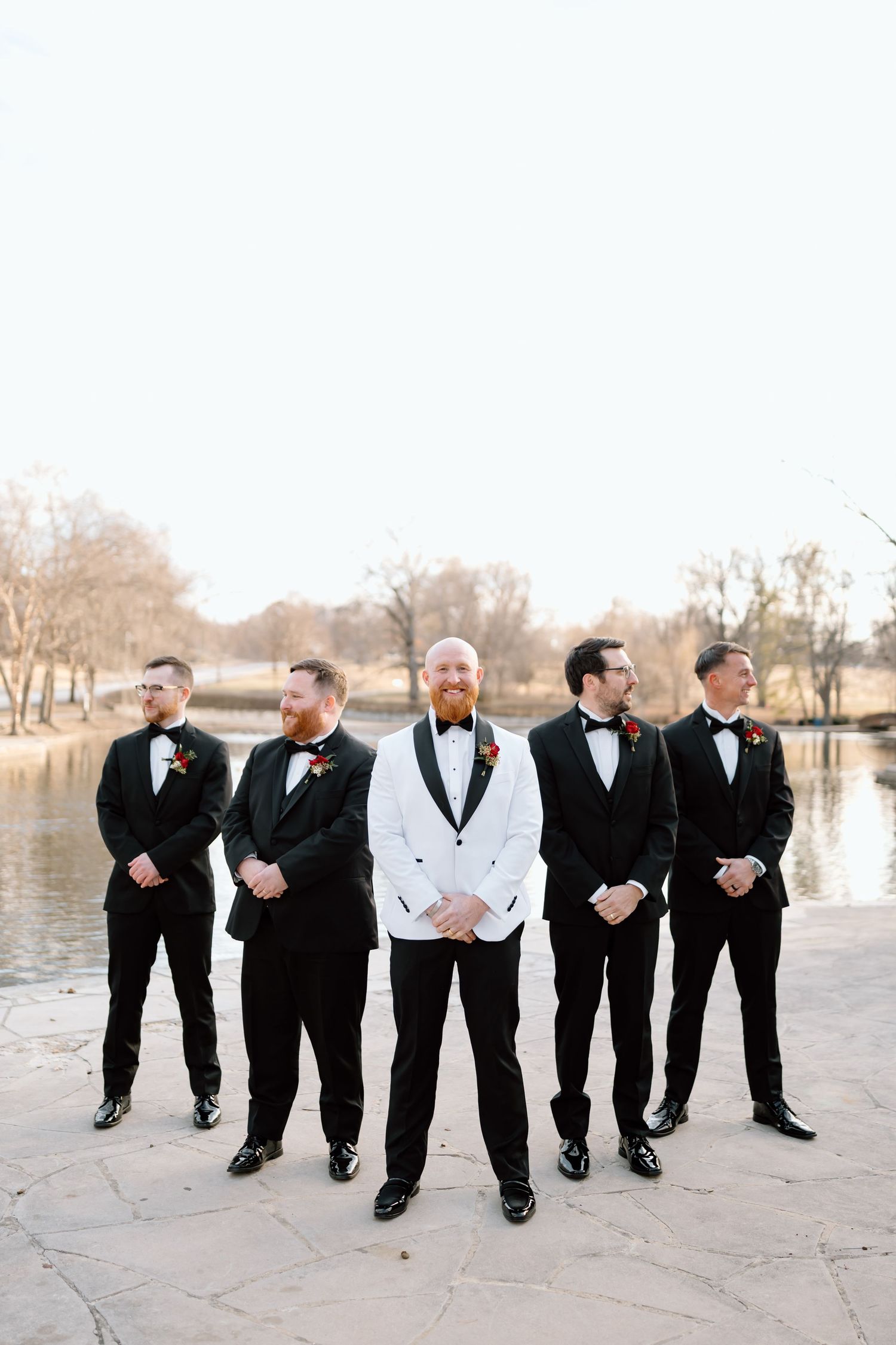 A group of groomsmen in black tuxedos and white shirts pose together by a lake on a winter day.