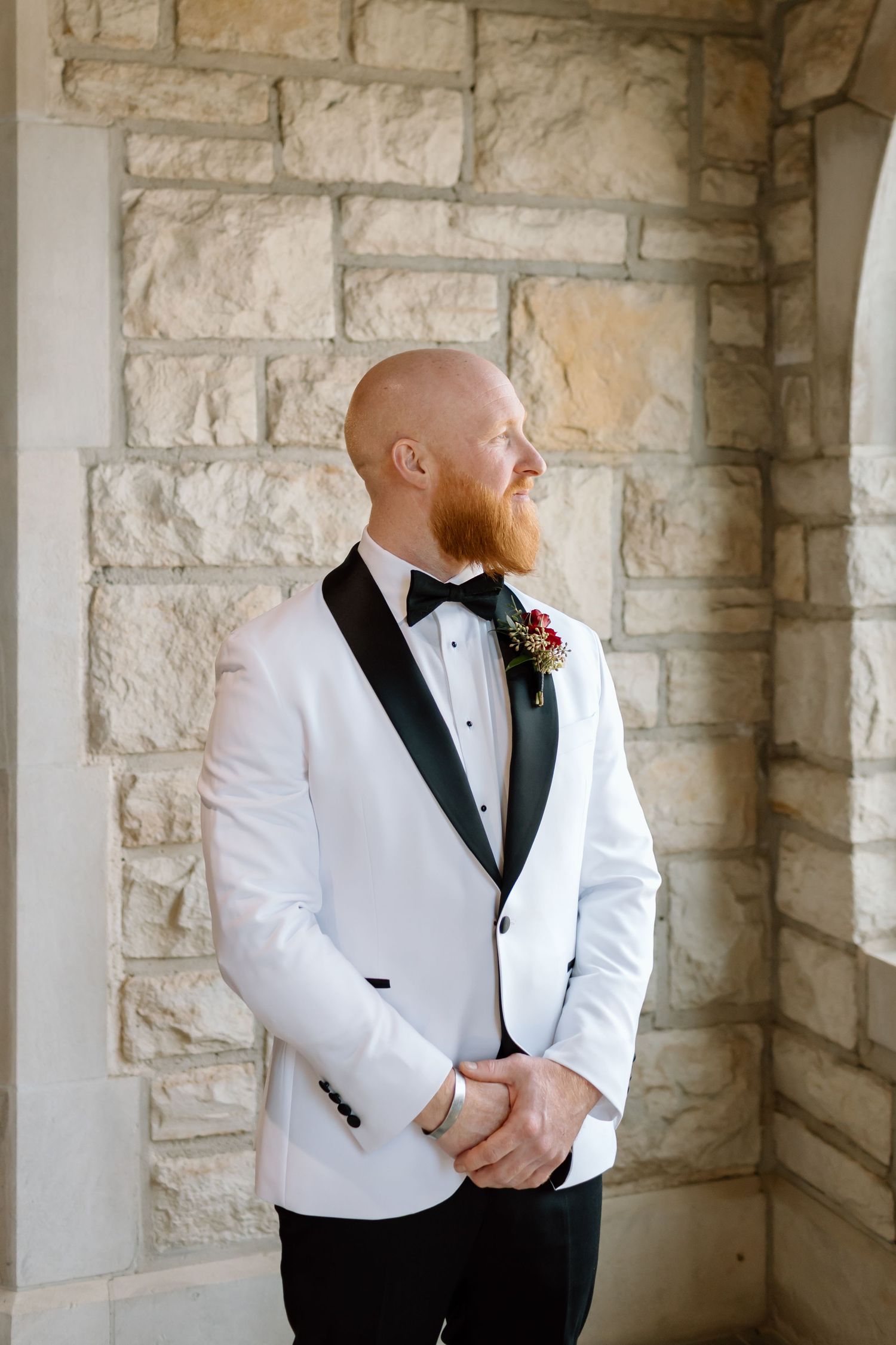 A groom in a white tuxedo jacket and black bow tie poses against a stone wall in an elegant setting.