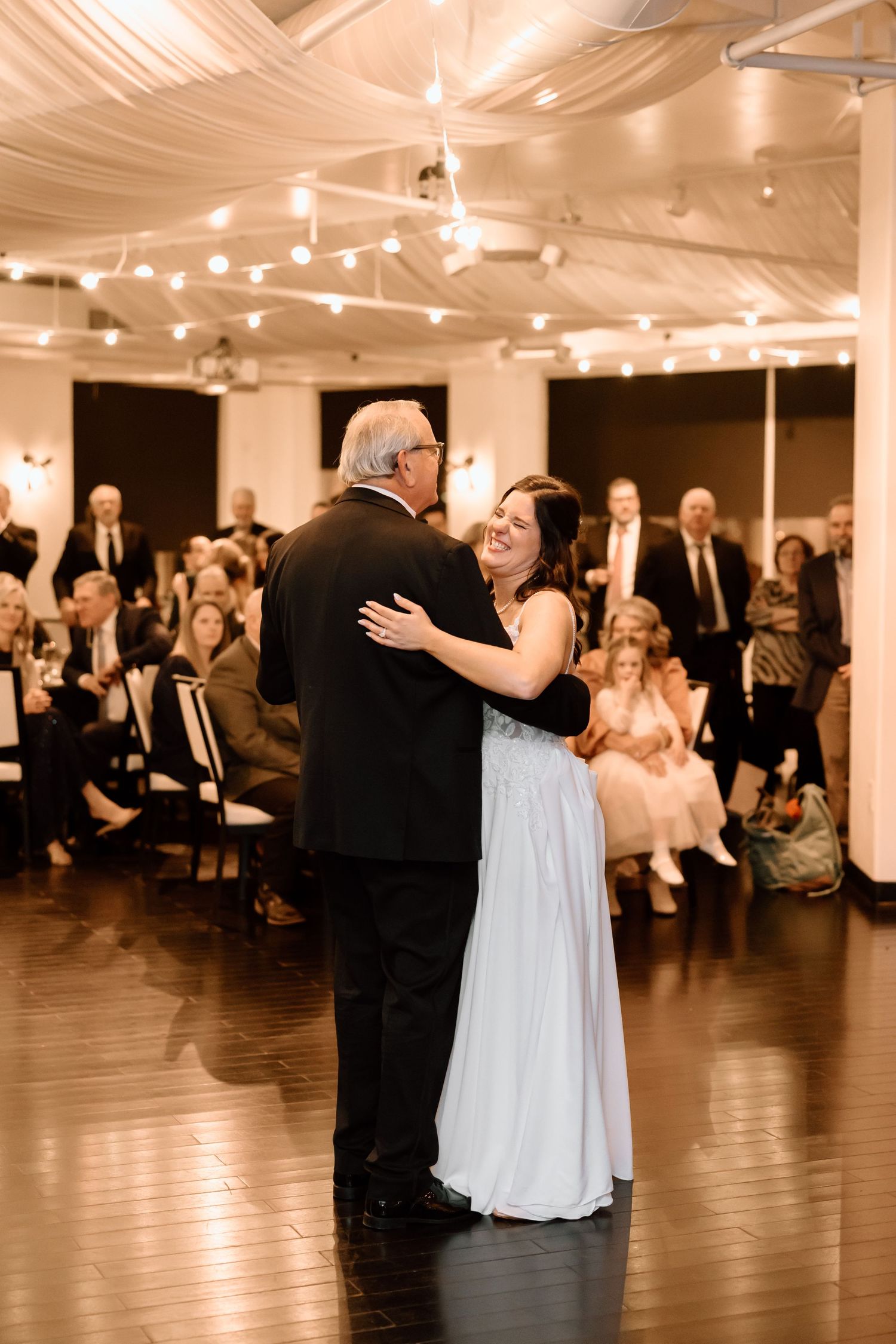 A bride and her dance partner share a special moment on the dance floor under warm lighting at an indoor wedding reception.