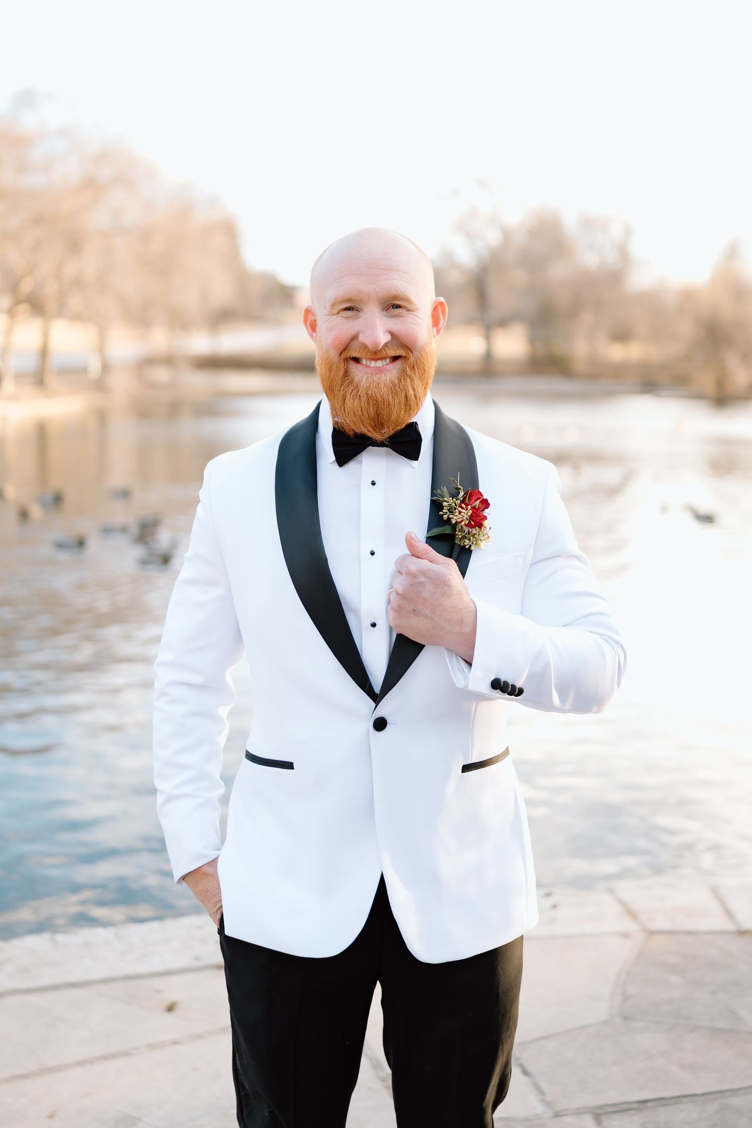 Groom in white tuxedo jacket and black bow tie poses by waterfront at sunset.