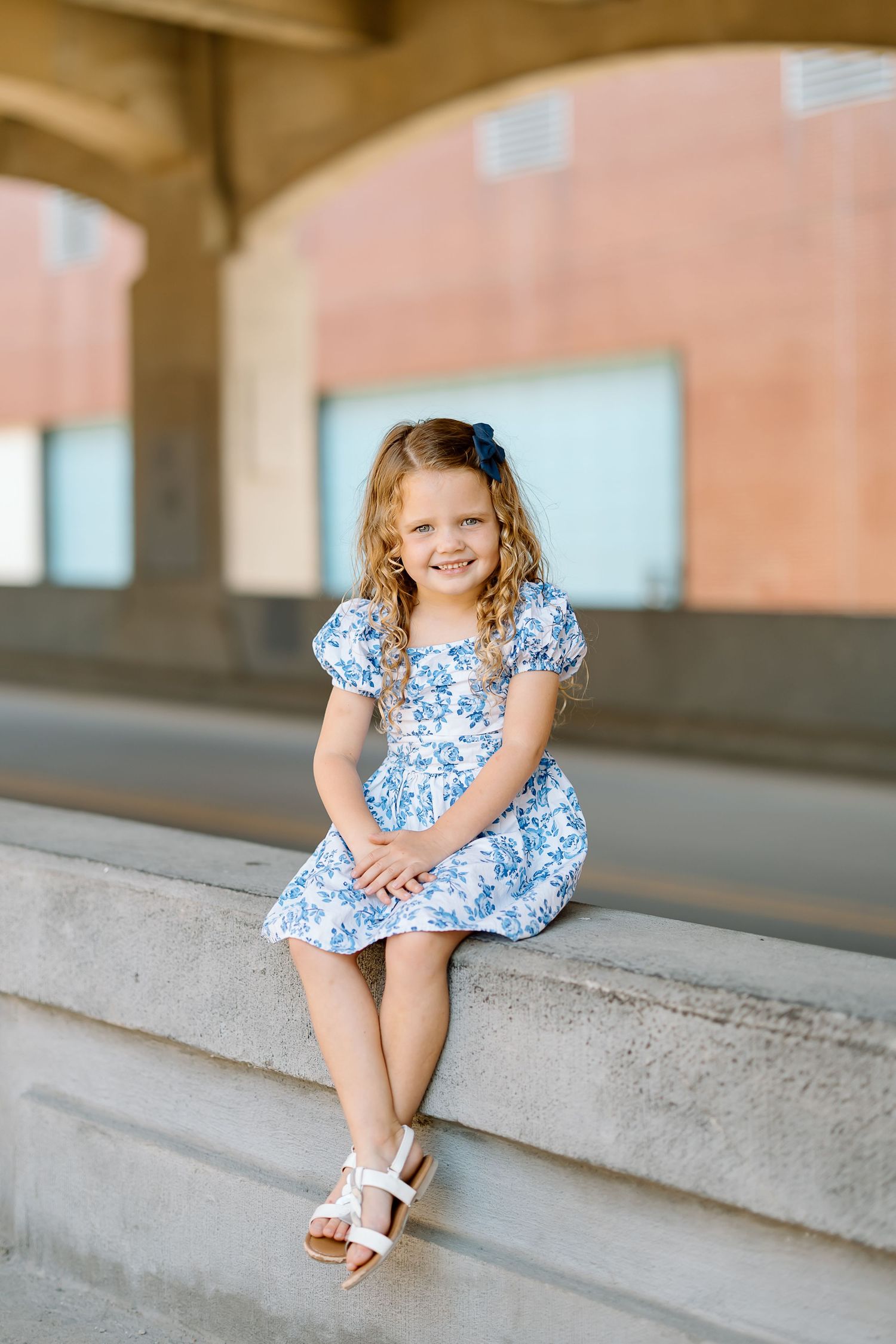 A young child in a blue floral dress sits on concrete steps wearing white sandals on a sunny day.
