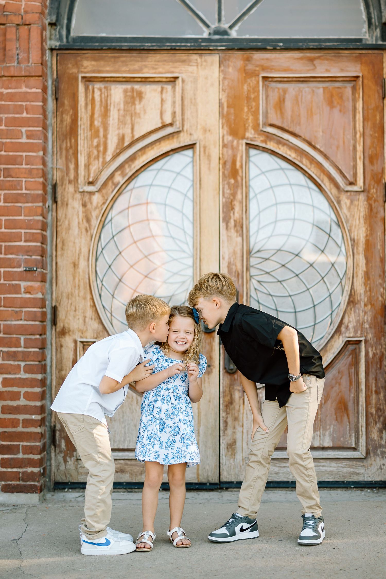 Three children share a playful moment in front of vintage wooden doors with circular glass panels and brick walls.