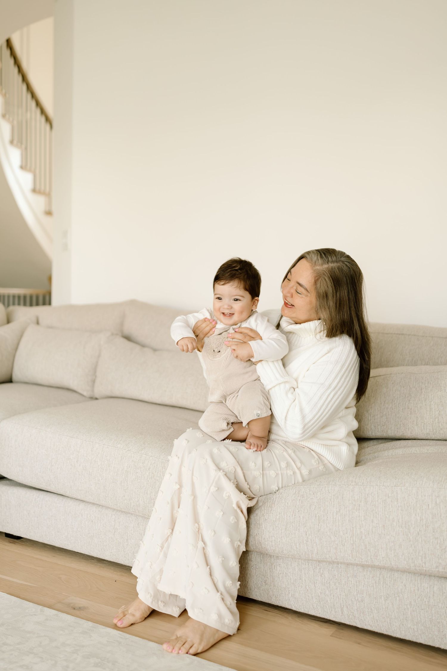 A tender moment shared between parent and baby on a light gray couch in a bright, minimalist living space.