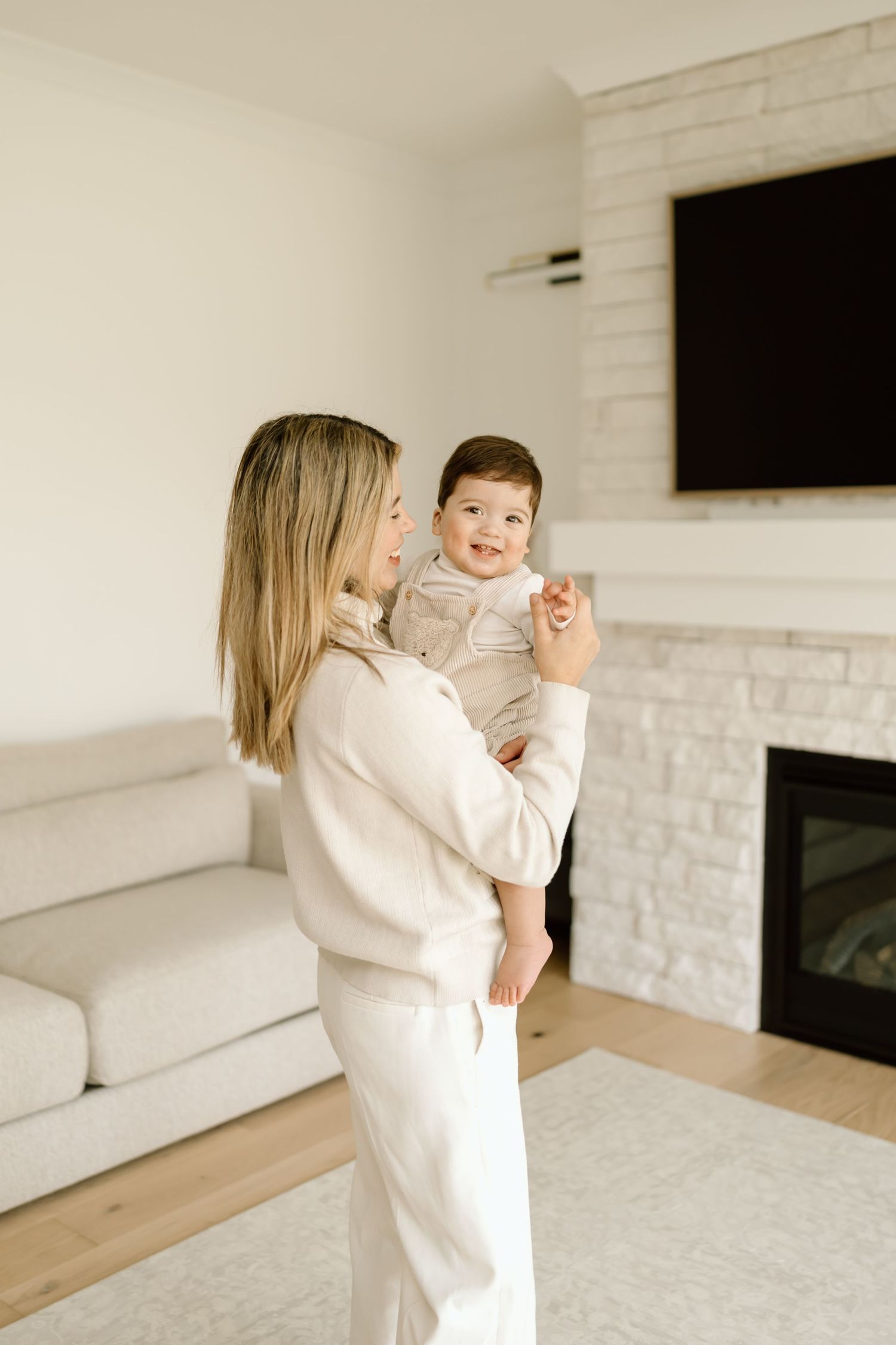 A parent in a white sweater holds and plays with a baby in a bright, modern living room with white brick walls.