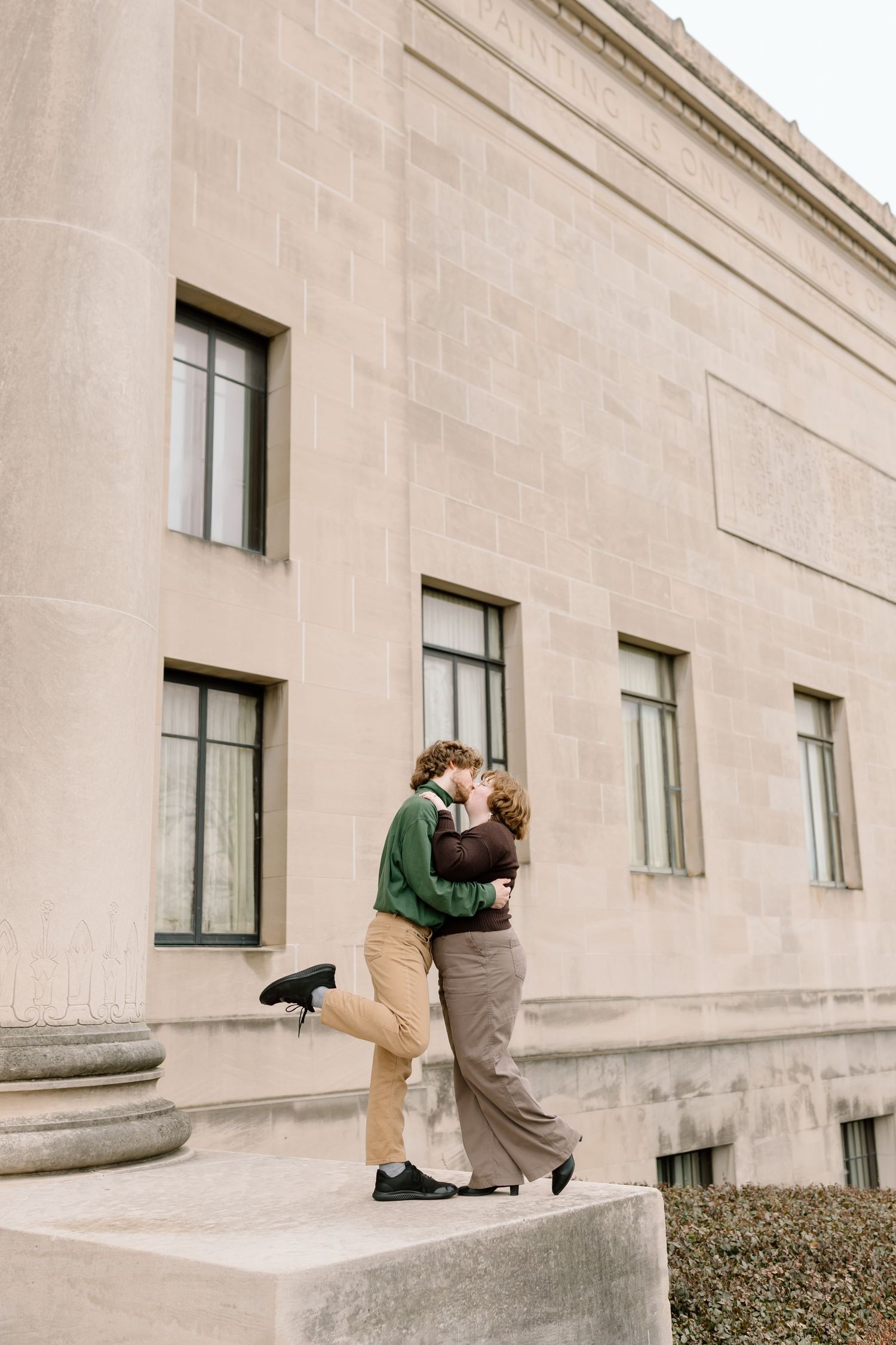 A couple shares a romantic kiss on stone steps outside a beige building with multiple windows.