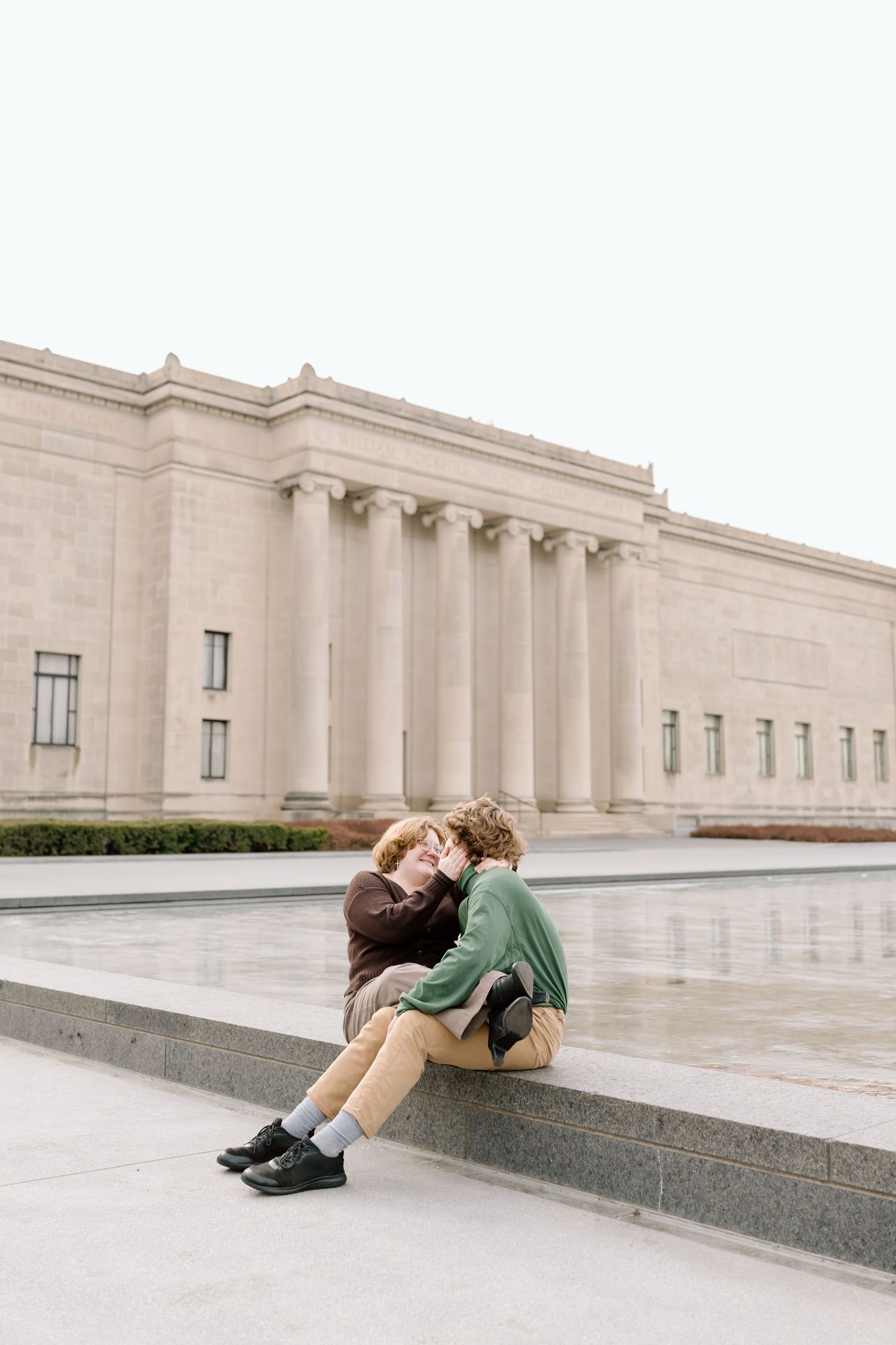 A person in a green sweater rests against a stone ledge outside a grand neoclassical building.