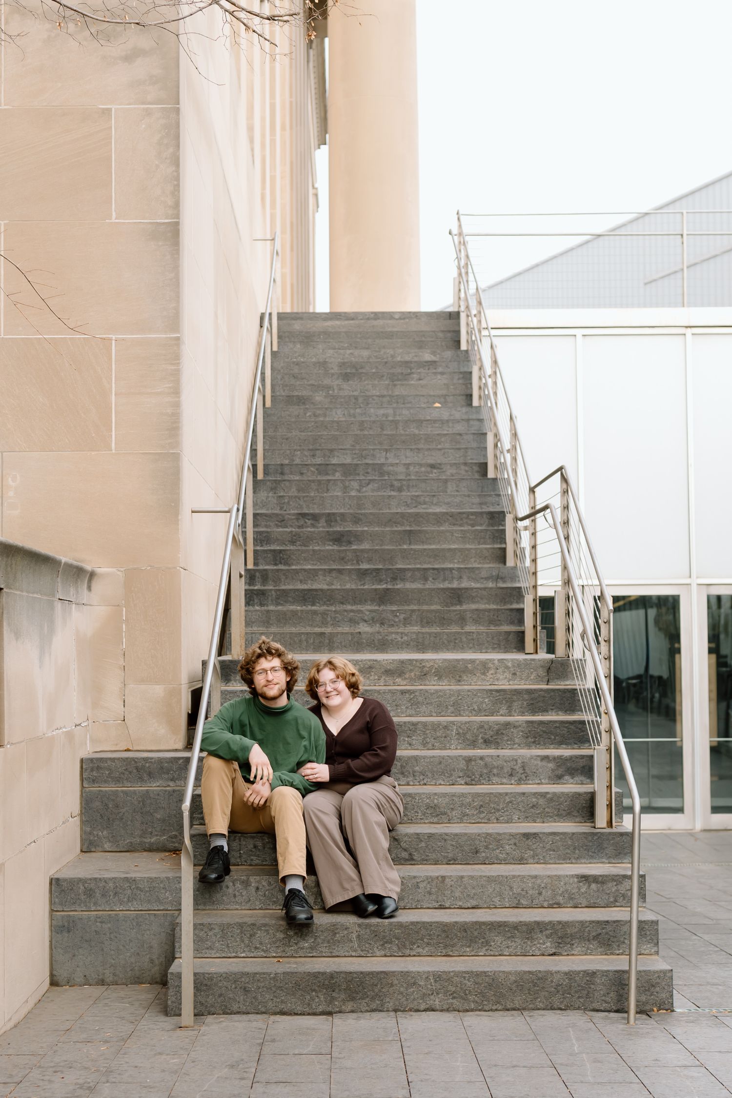 Two people sit together on concrete steps outside a modern building with beige walls and metal railings.