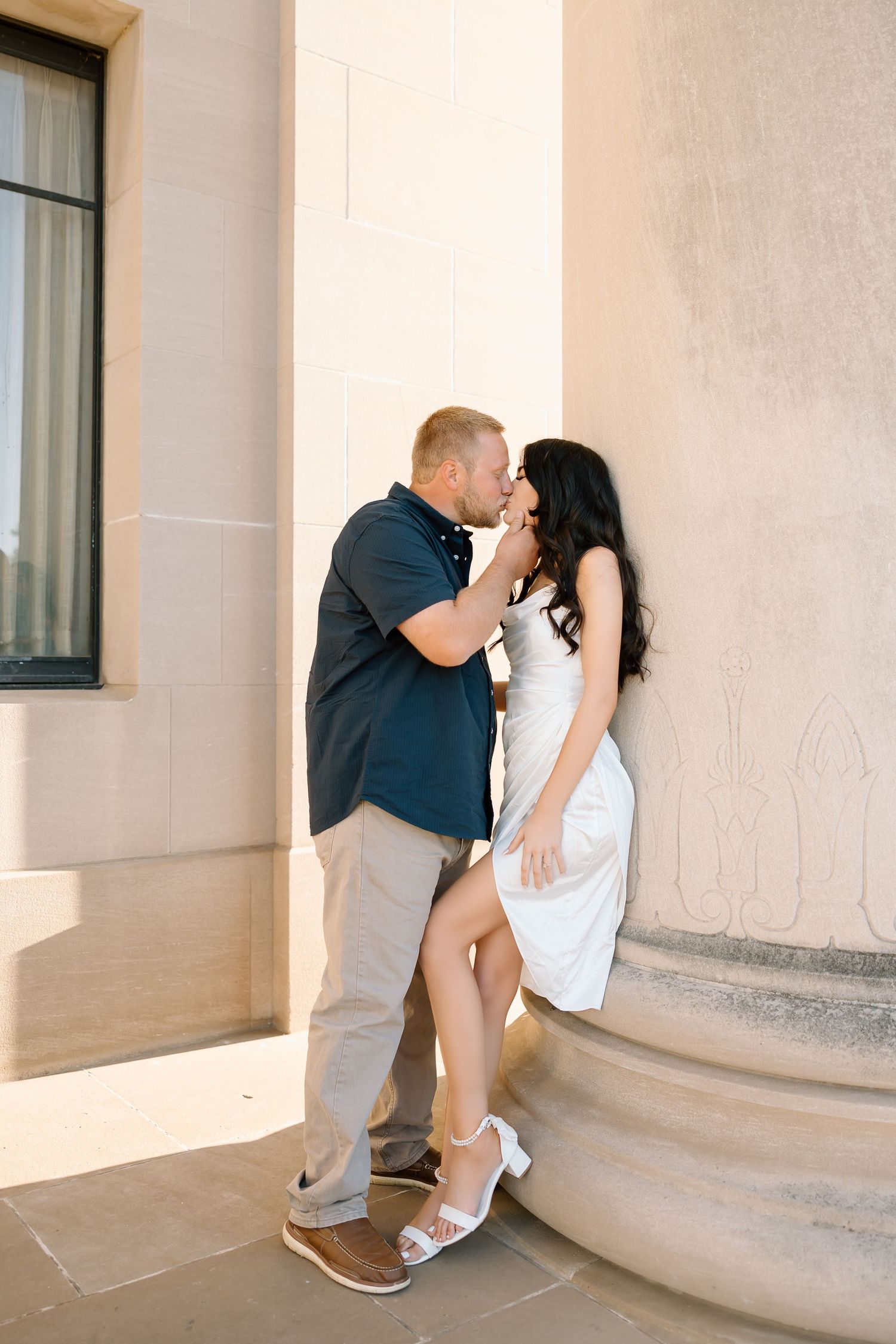 A romantic engagement photo series showing a couple in casual attire against a light colored building wall.