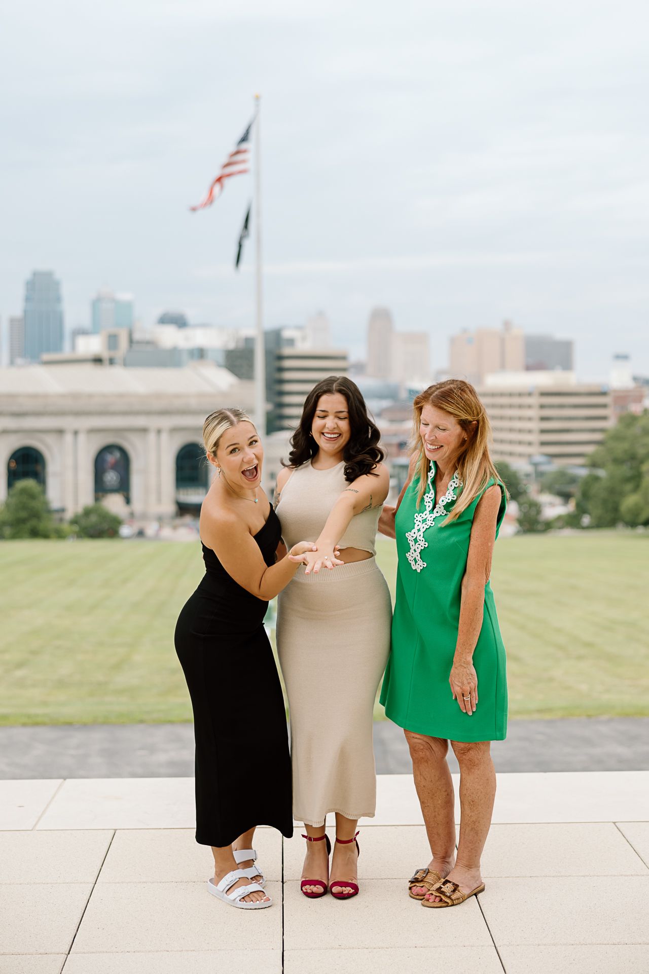 Three friends in summer dresses pose together on a concrete plaza with city skyline behind them.