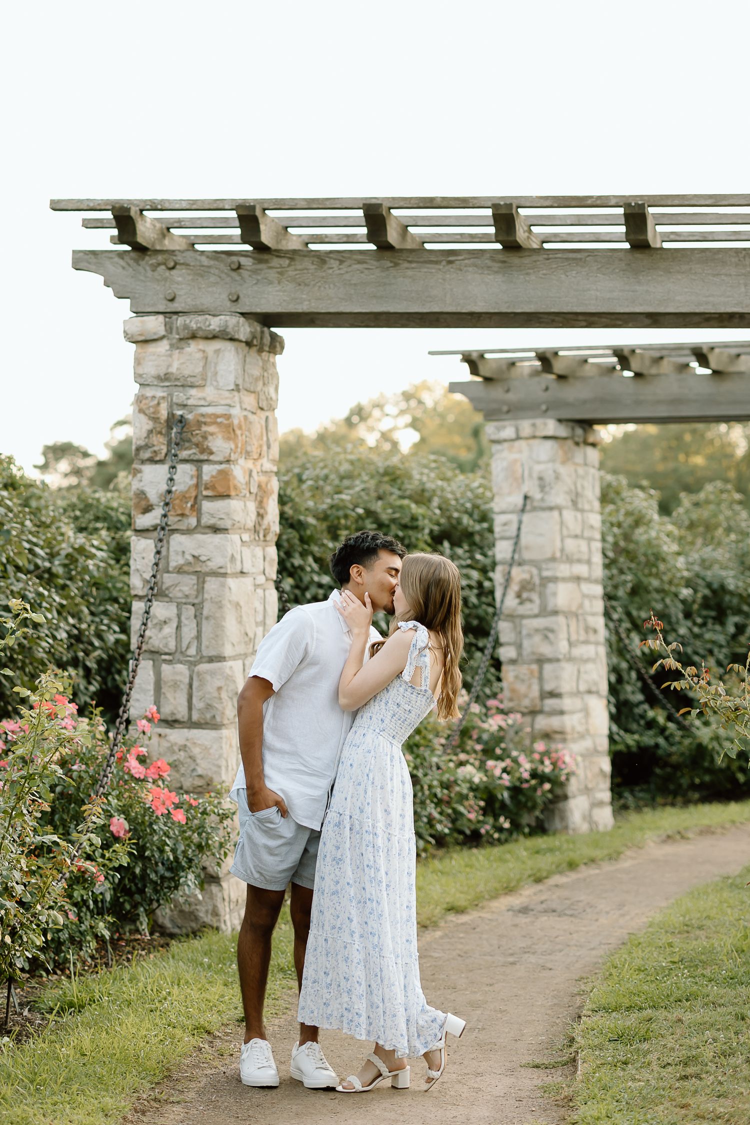 A romantic couple shares tender moments under a stone pergola surrounded by blooming roses in a garden setting.