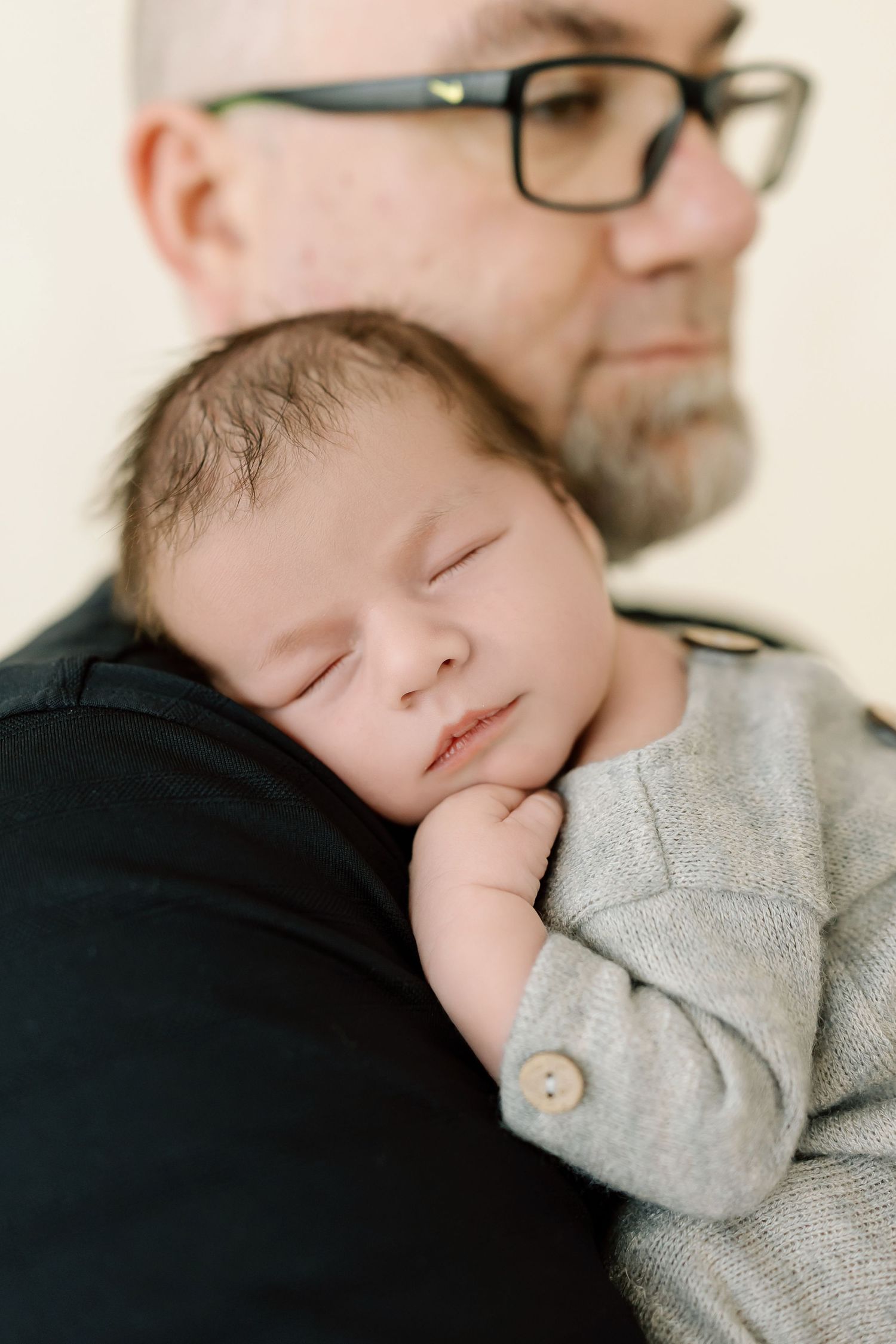 A sleeping newborn baby in a gray sweater rests peacefully on someone's shoulder wearing black.
