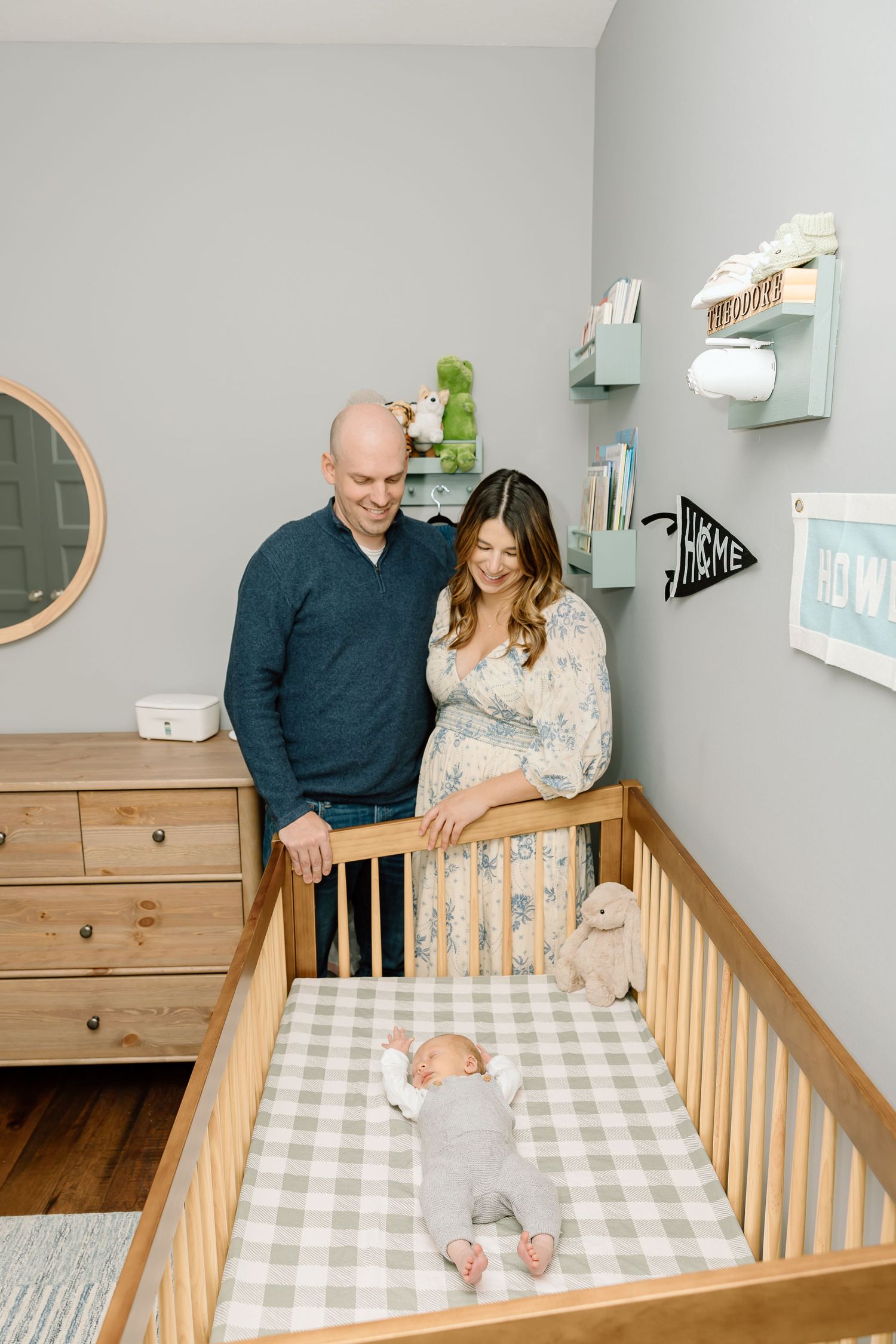 Parents lovingly stand over wooden crib with buffalo check bedding in modern nursery setting.