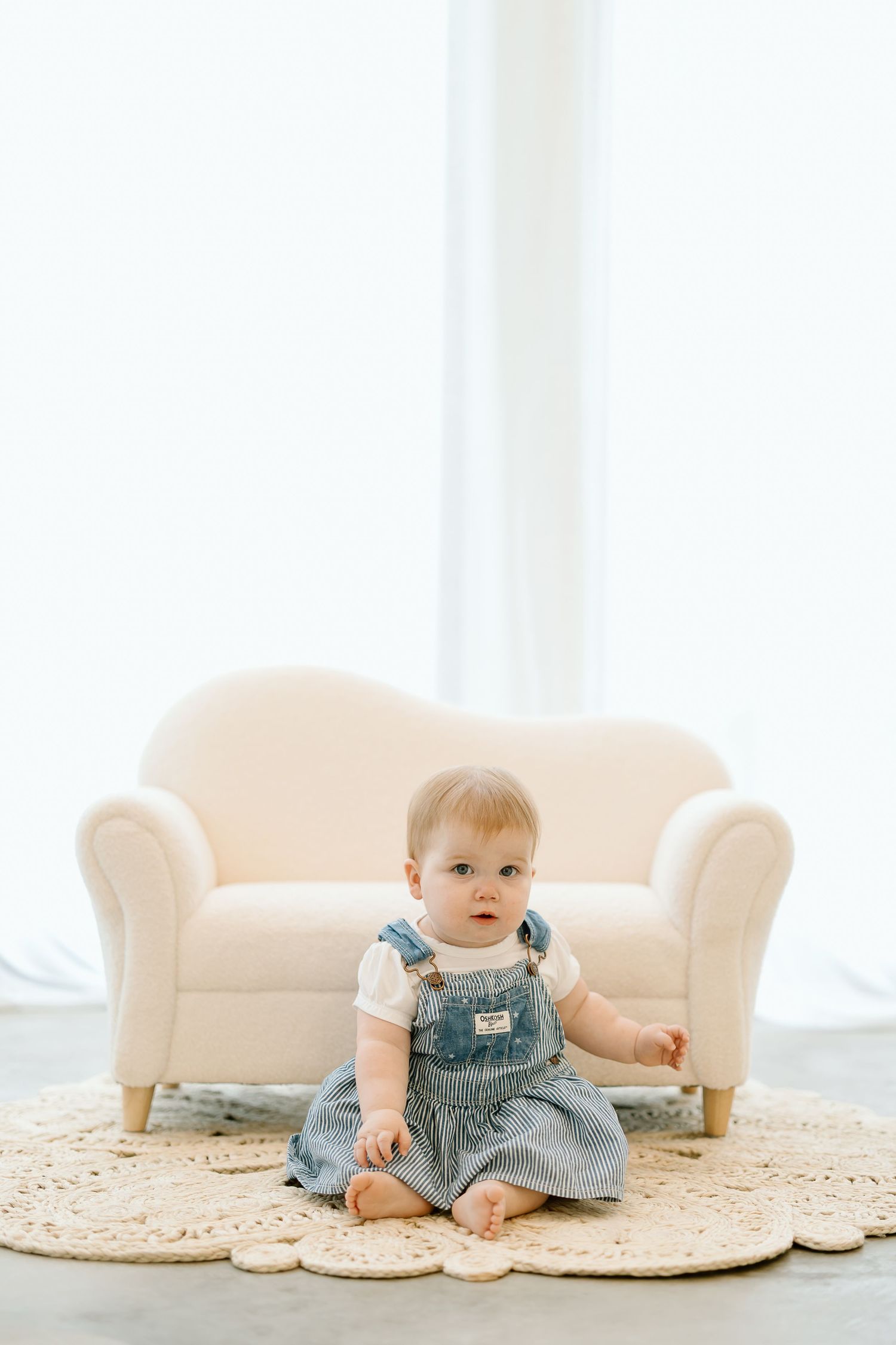 Baby in denim overalls sits on a round jute rug in a bright, minimalist room with cream-colored furniture.