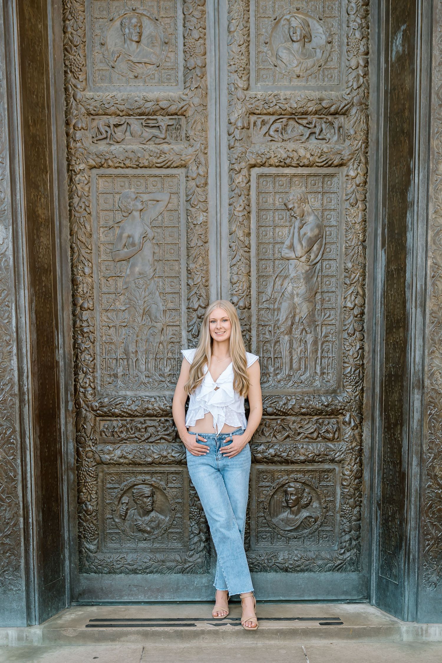 A person in light jeans and white top poses against ornate carved metal doors with decorative panels.