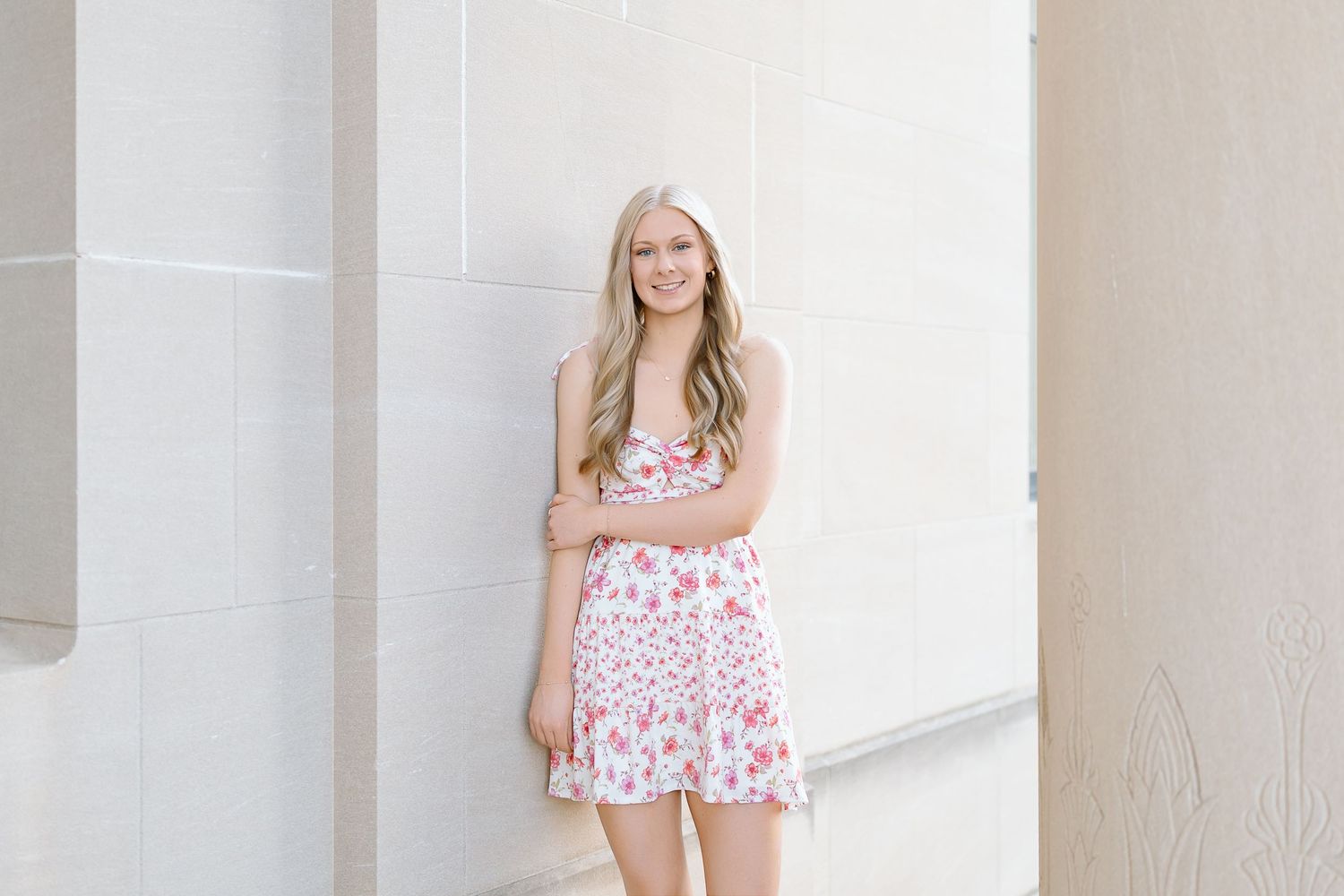 A person in a pink floral summer dress stands smiling against a white column background.