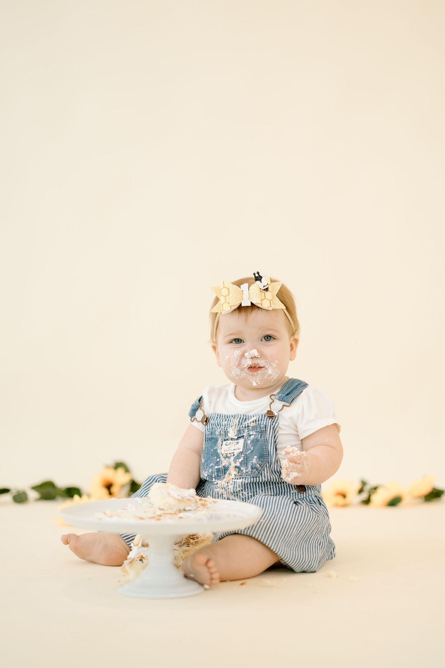 A small child in blue and white striped overalls sits on a cream background with a cake and flower crown.