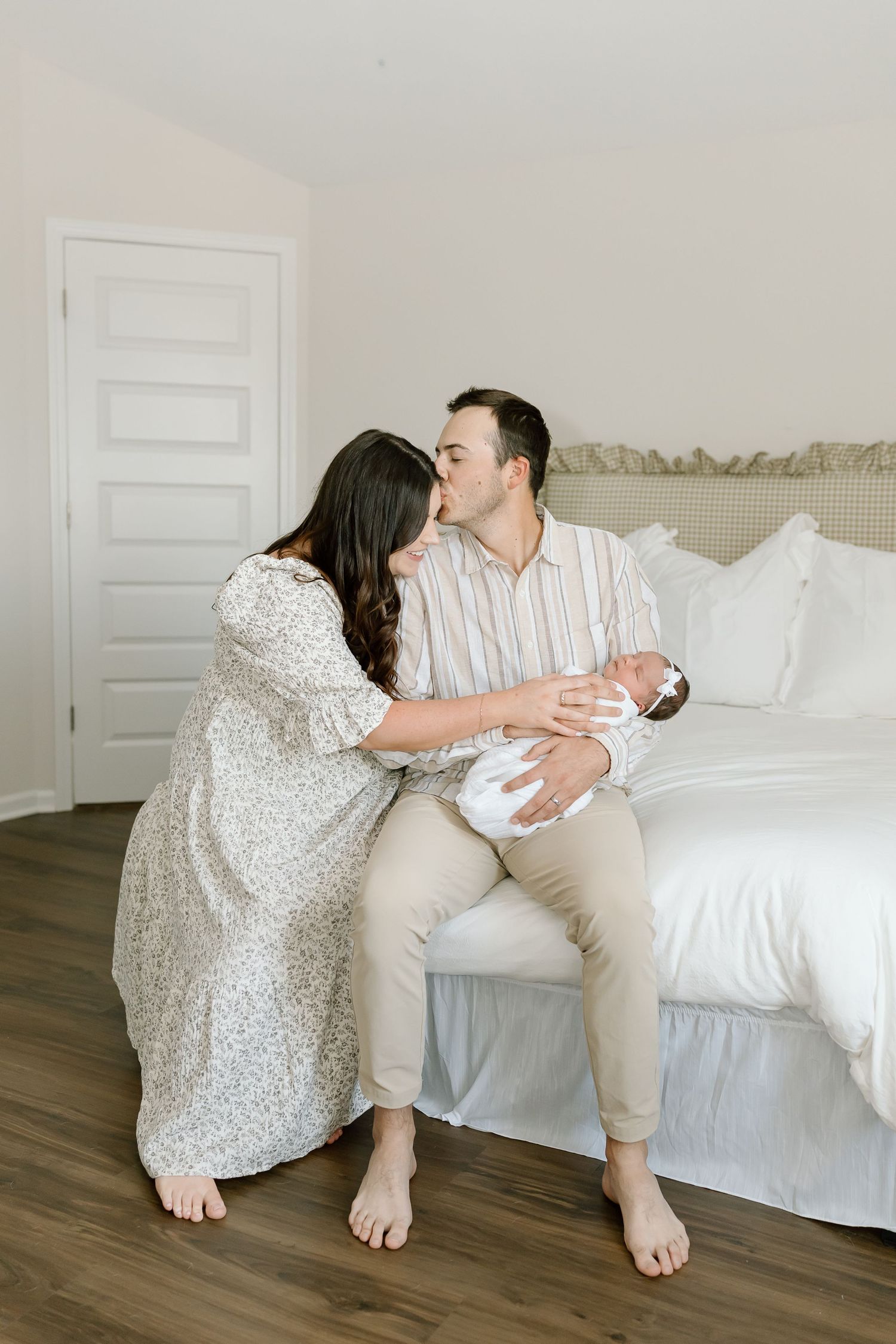 A couple sits on a white bed in a bright room, sharing an intimate moment while holding their newborn baby.