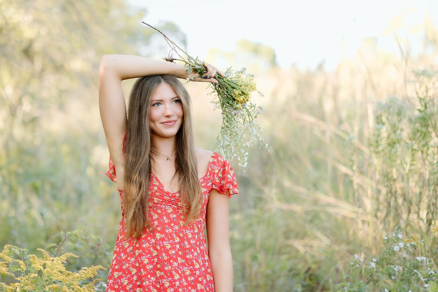 A woman in a red floral dress poses outdoors in a sunlit meadow with tall grass and wildflowers.