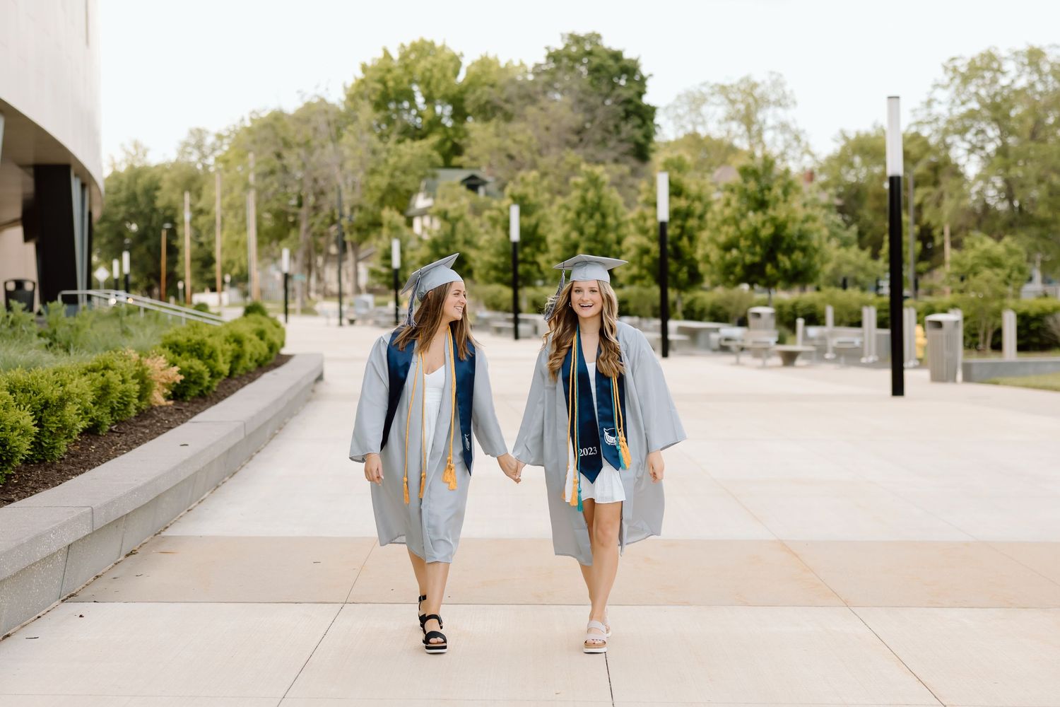 Two graduates in matching light blue gowns walk hand in hand on a sunny campus pathway.