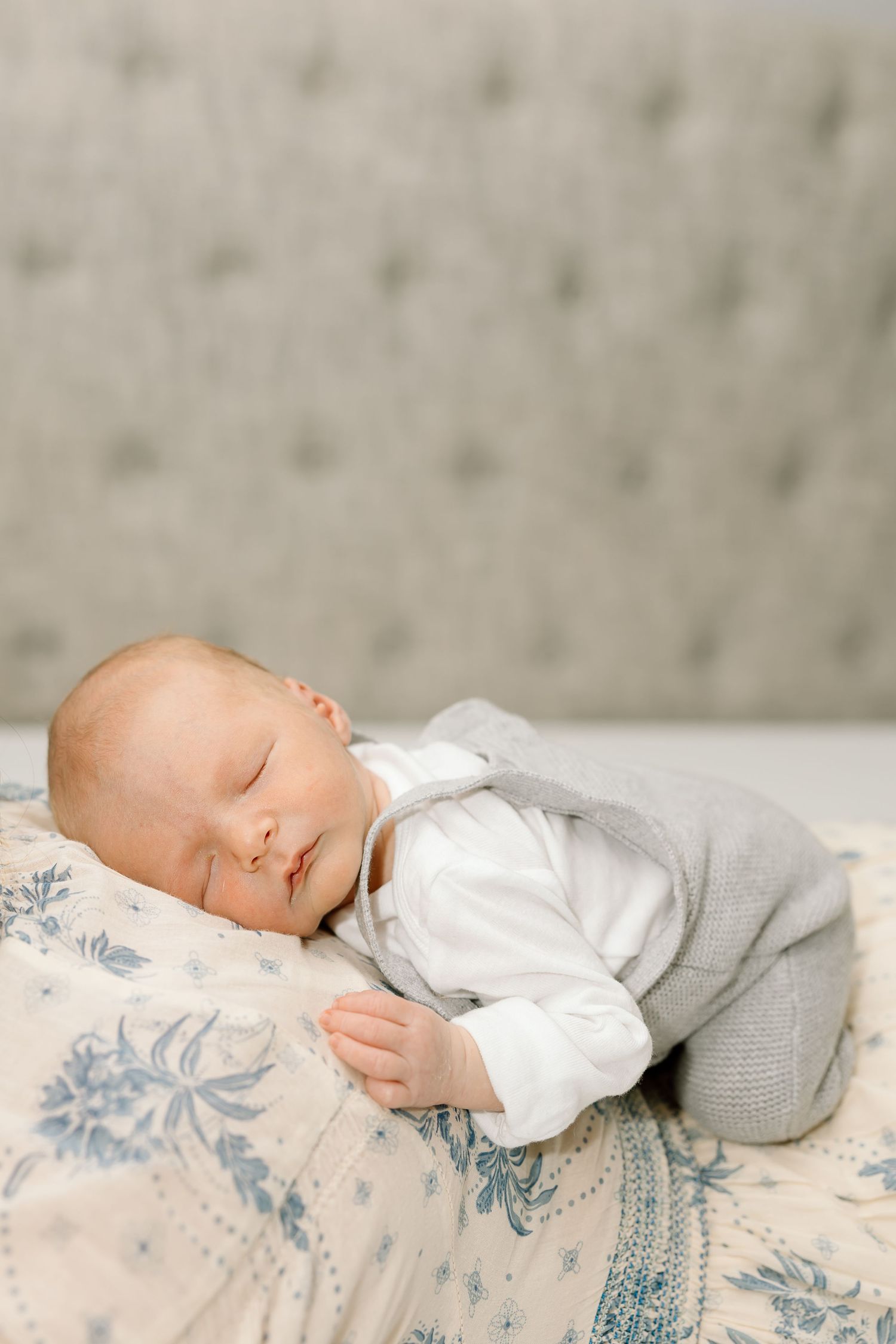 A sleeping baby wearing white and gray clothing rests peacefully on a blue floral patterned surface.
