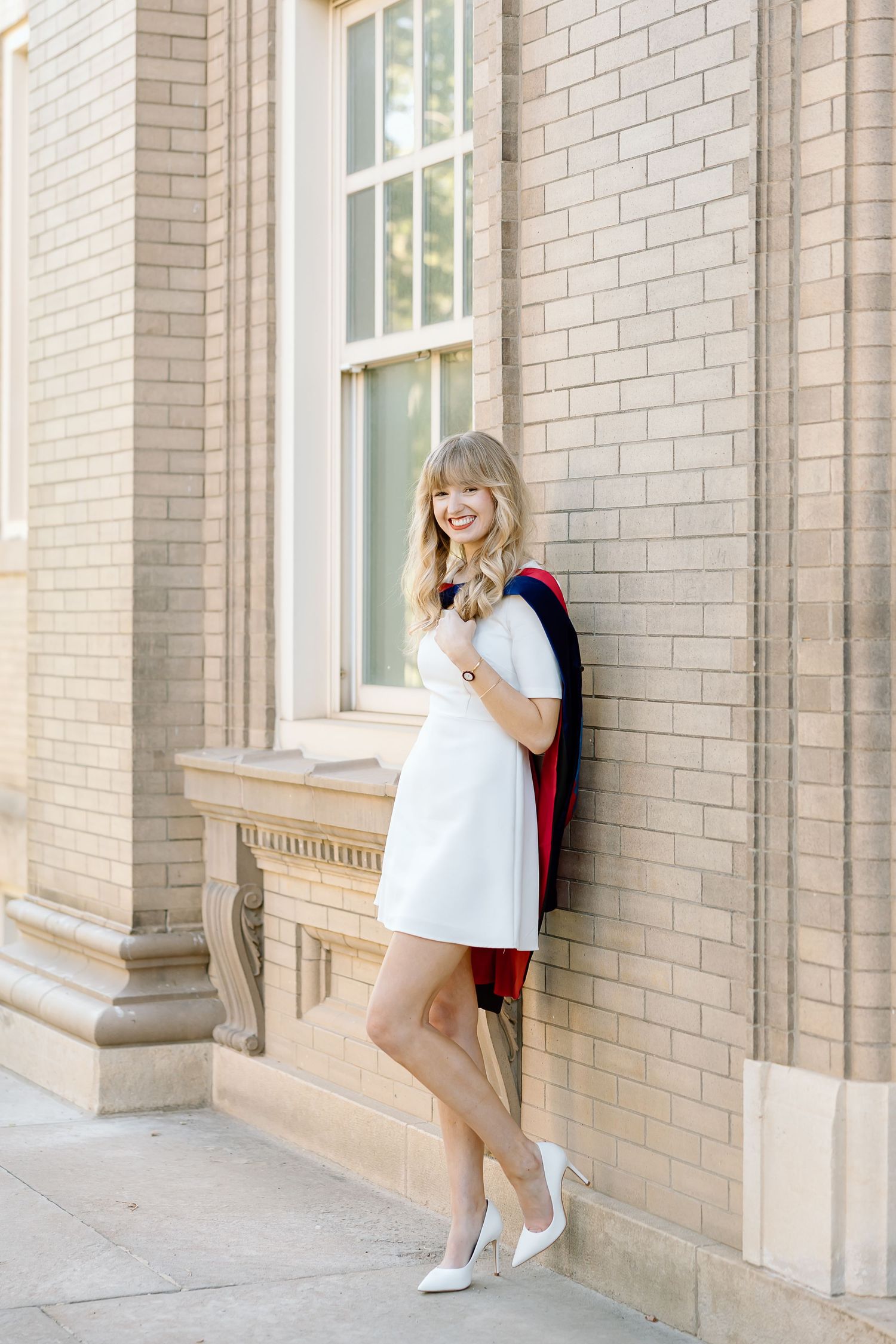 A person in a white dress and sneakers poses playfully against a light brick wall near large windows.